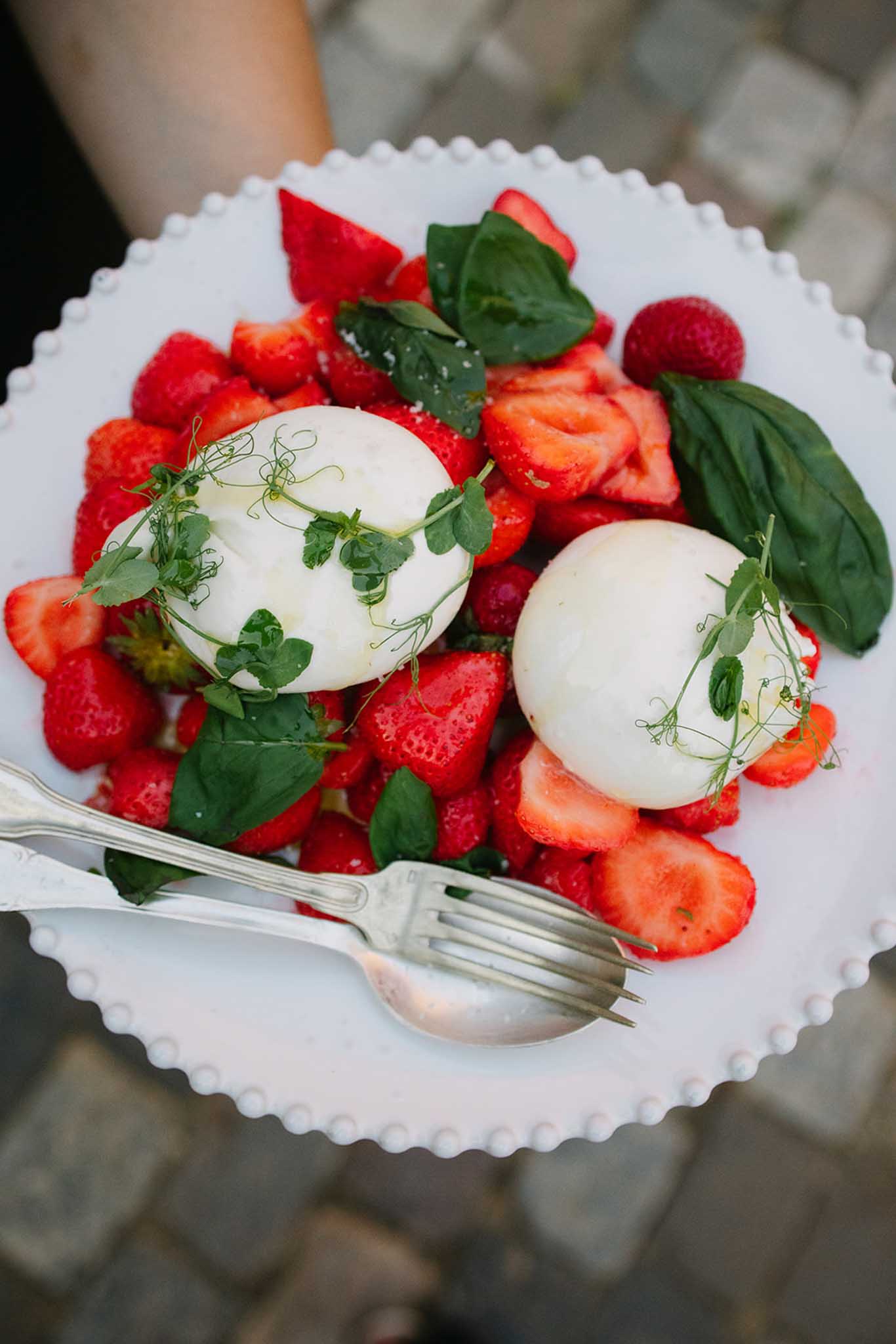 Plate of burrata and fresh strawberries garnished with microgreens and basil, held by guest during reception