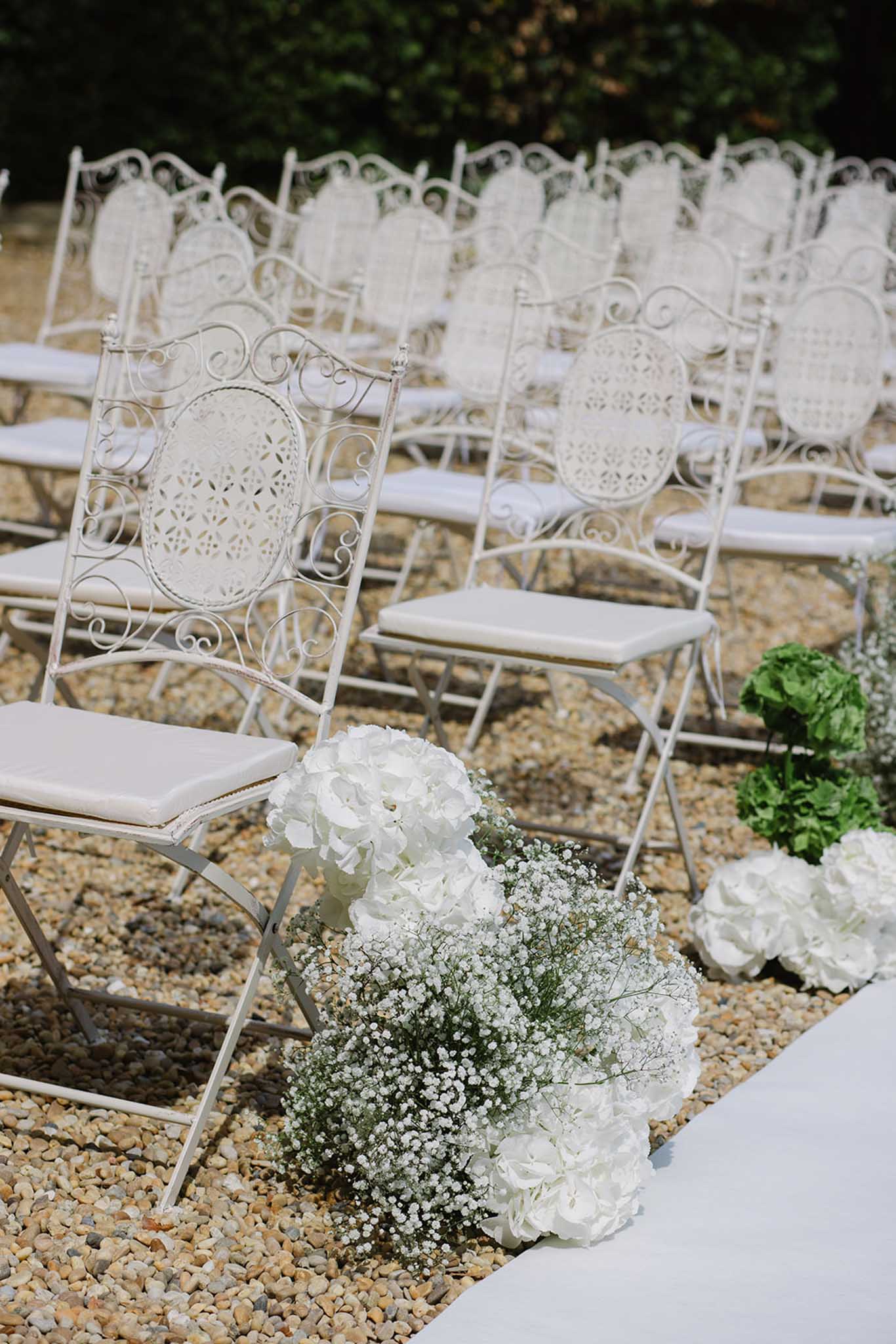 Ceremony aisle with ornate white metal chairs and white hydrangea and baby's breath aisle arrangements on gravel path