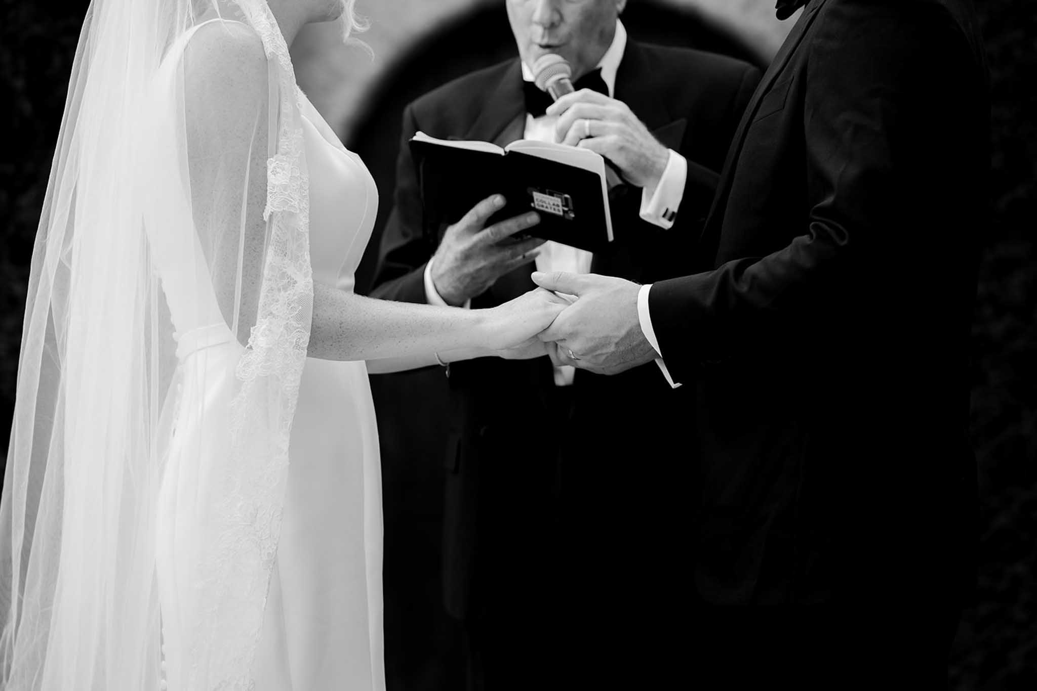 Black and white close-up of couple exchanging rings with officiant reading during ceremony