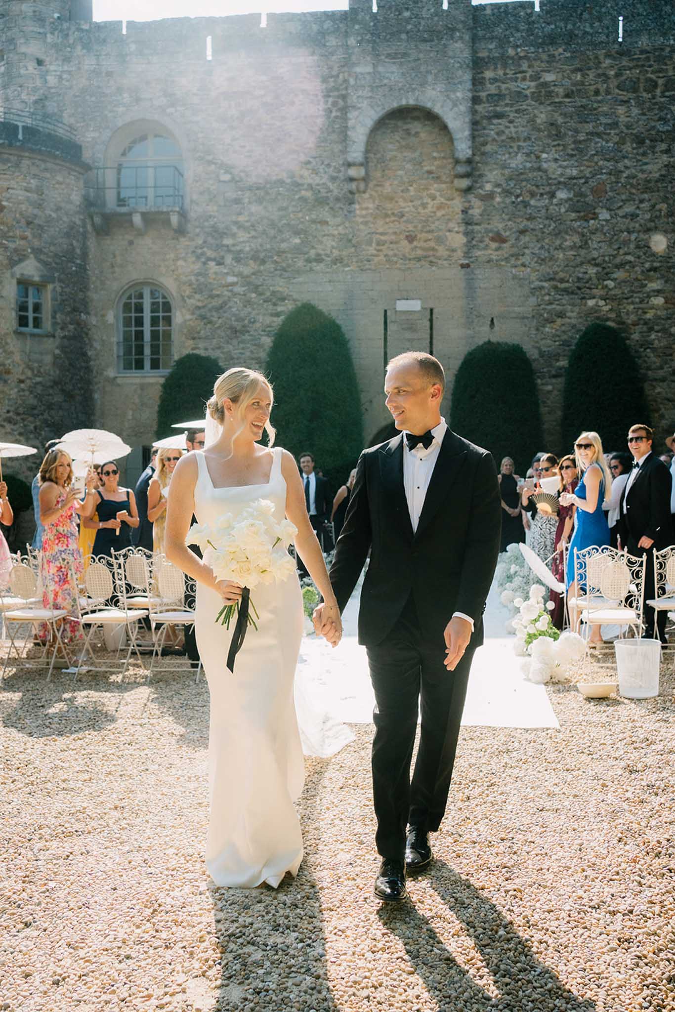 Bride and groom walking after ceremony at ivy-covered stone castle with guests seated on white chairs on both sides
