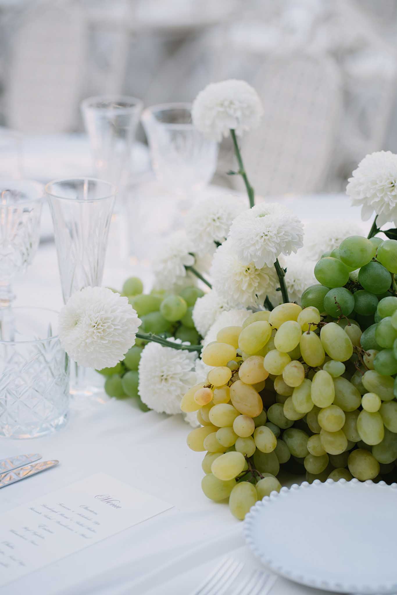 White dahlia and cascading green grape centerpiece on white linen table with glassware and handwritten place cards