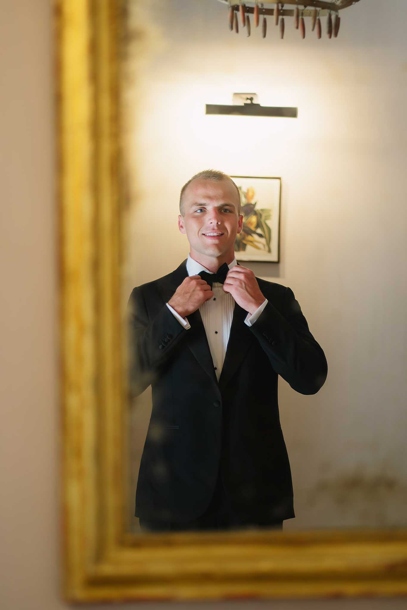 Groom adjusting black bow tie and smiling in cream-walled room during wedding preparations
