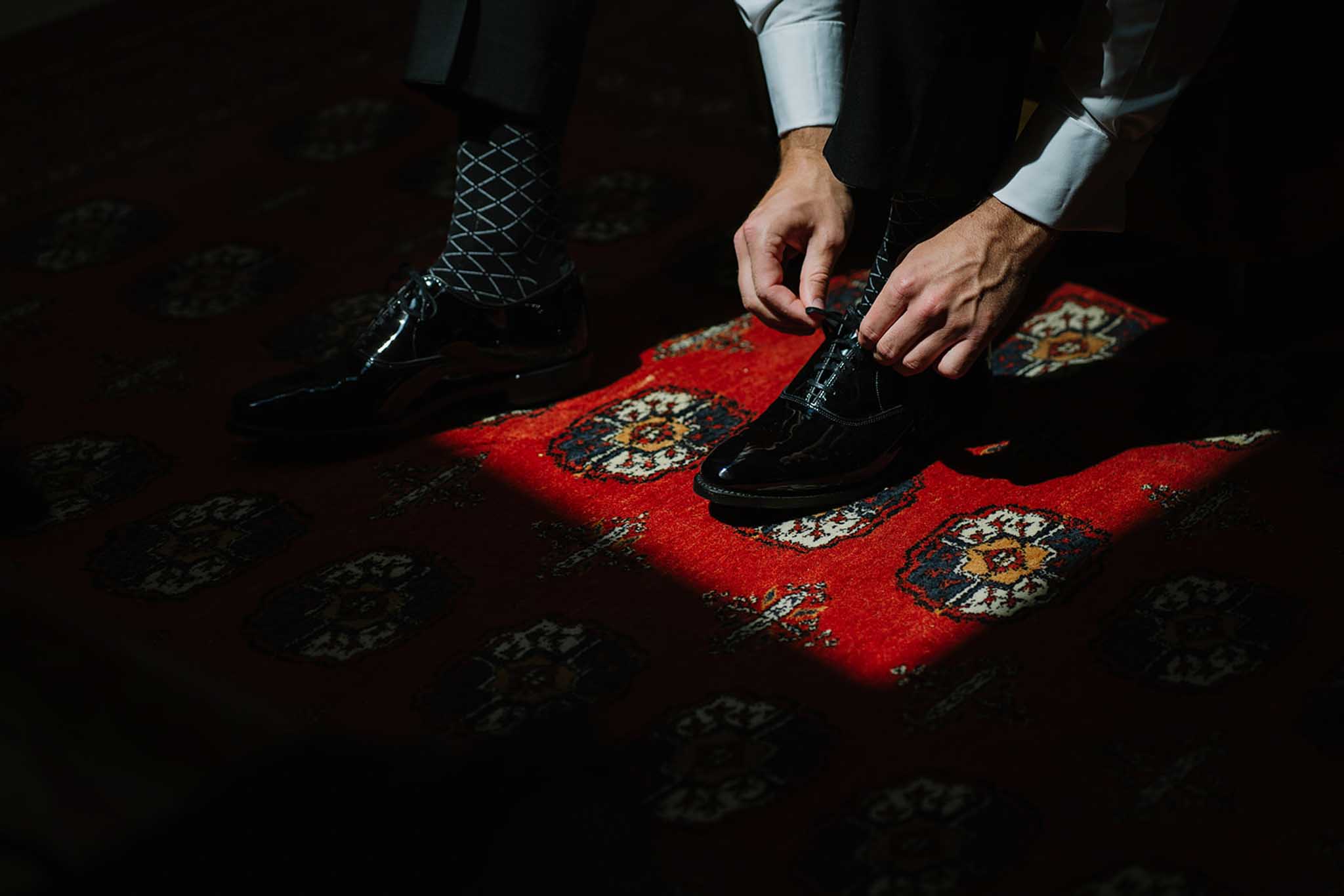 Two grooms adjusting black patent leather shoes on ornate red and gold carpet runner, light blue shirts visible