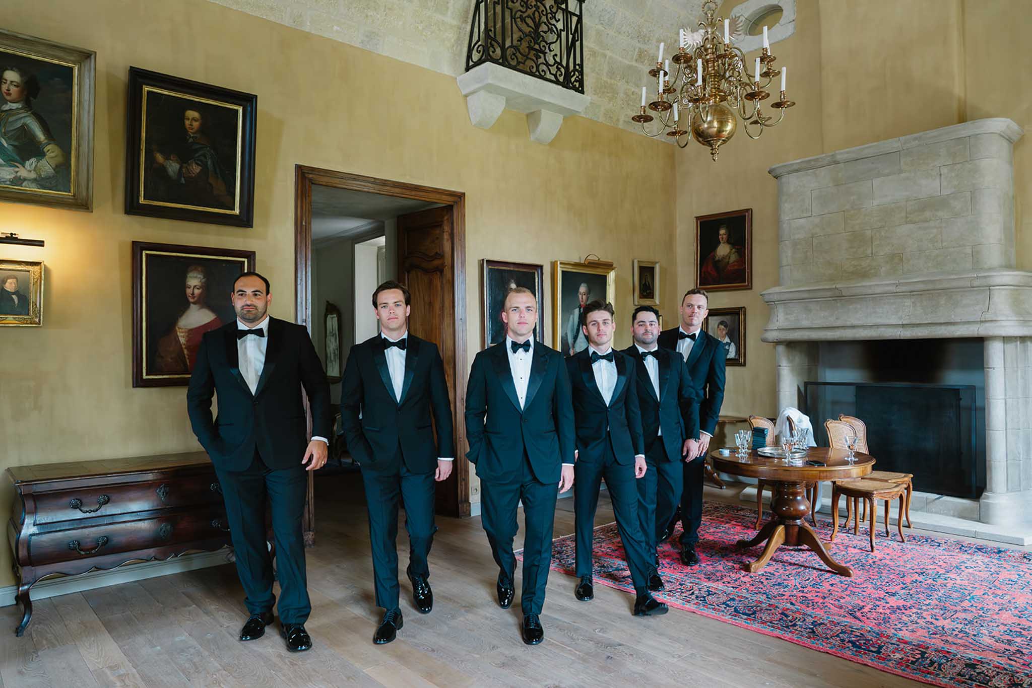 Groom and six groomsmen in navy tuxedos walk through grand ballroom with chandelier and fireplace