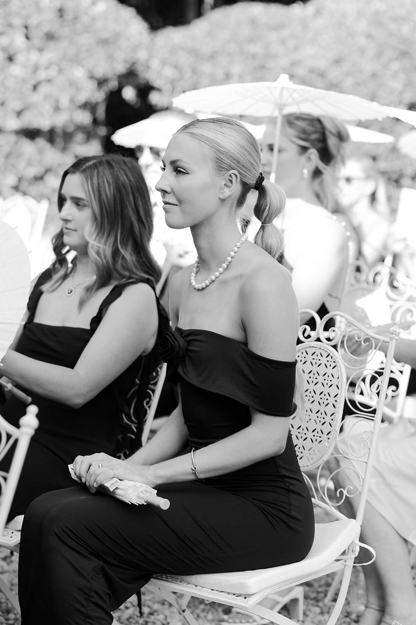 Black and white photo of formally dressed guests seated on white ornamental chairs during outdoor wedding ceremony