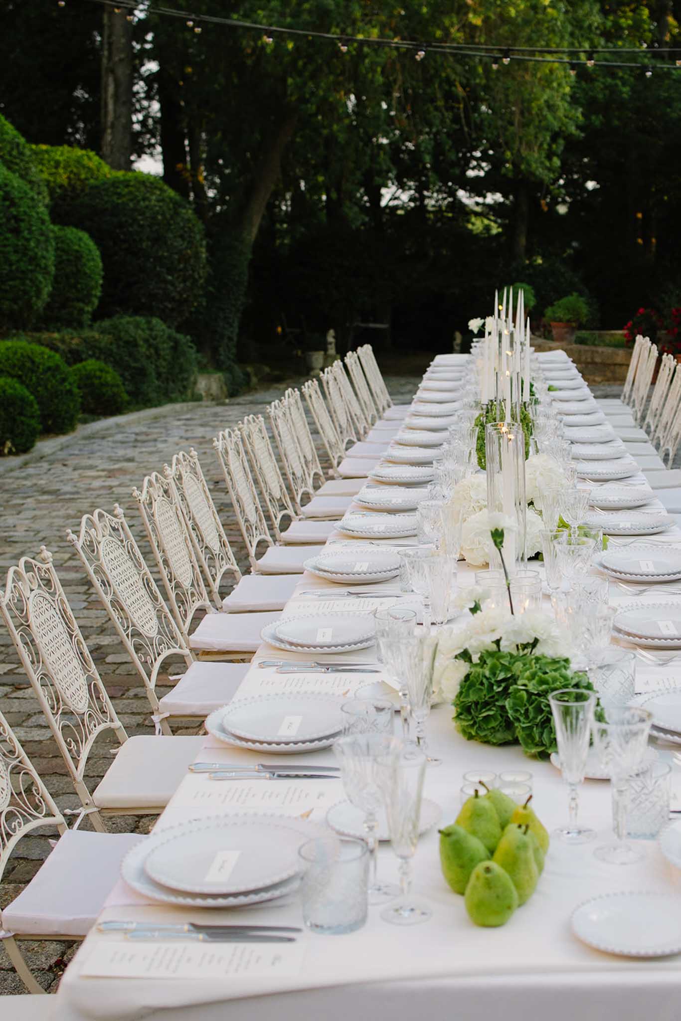 Long banquet table with pale pink linens, white chairs, and green pear accents beneath fairy lights in garden