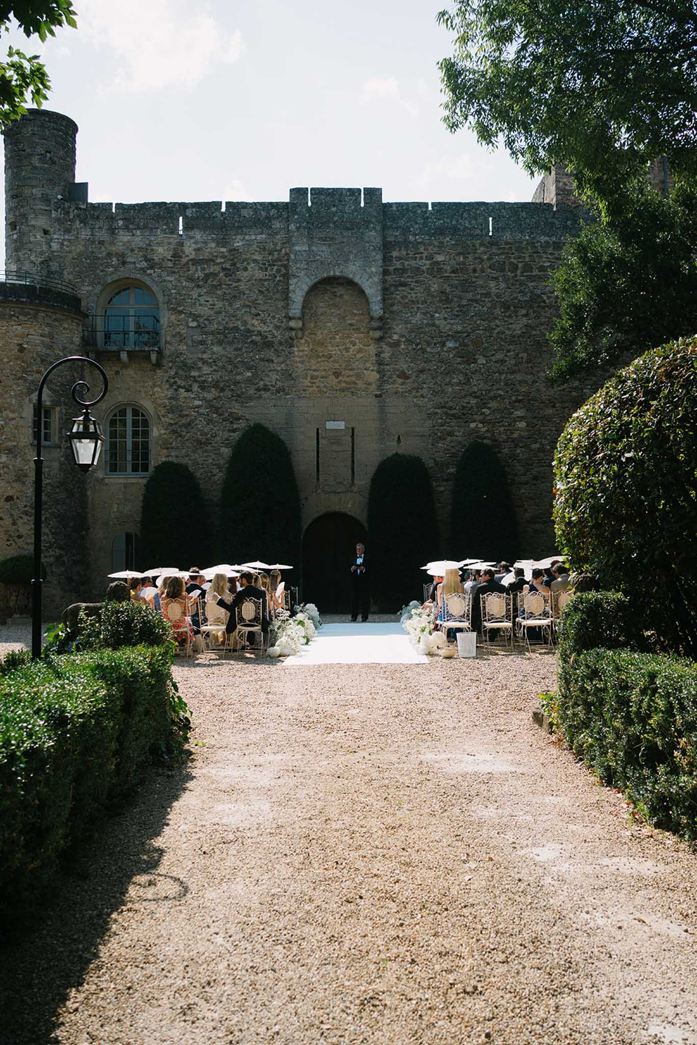 Wedding ceremony in medieval stone castle courtyard, gold Chiavari chairs, white umbrellas, crenellated walls as backdrop