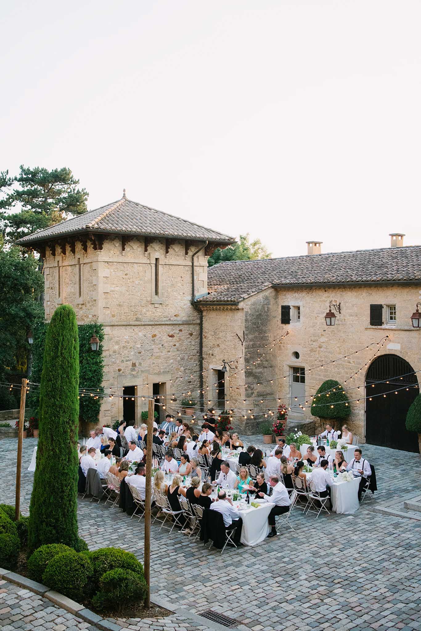 Wedding reception in a historic stone château courtyard with round tables, string lights and cylindrical cypress trees; circular tower on left.