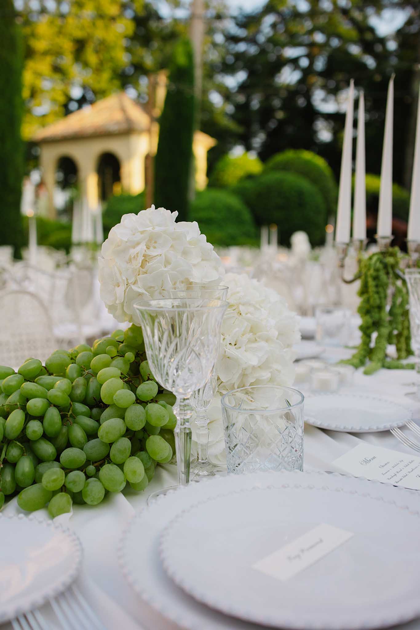 Reception table detail with green grapes, cream peony centerpiece, crystal glassware, and classical garden pavilion