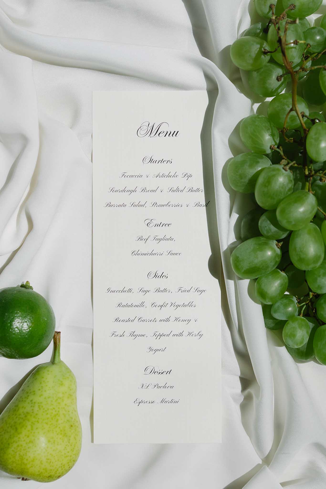 Wedding menu card on white linen styled with green grapes and pale pear as table decoration accents