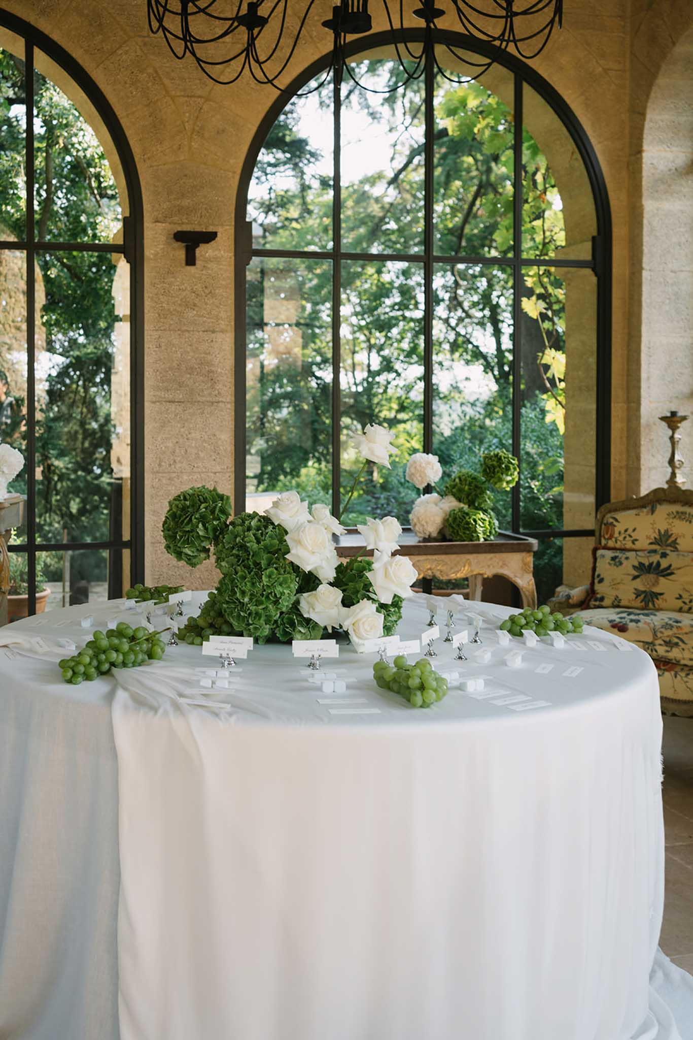 Round reception table with ivory linens, white rose and hydrangea centerpiece with grapes in a stone loggia with arched windows