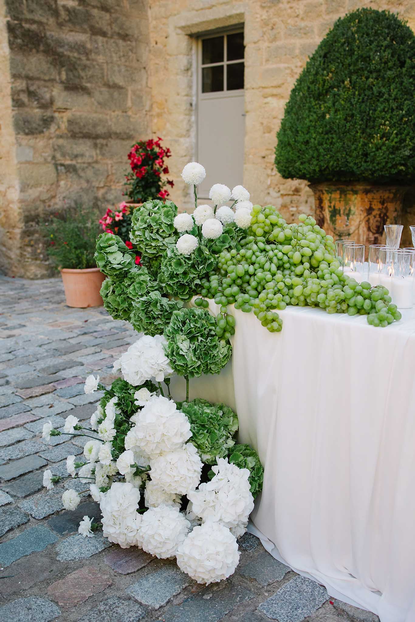White hydrangeas, green grapes, and foliage table arrangement on ivory linen at stone courtyard venue