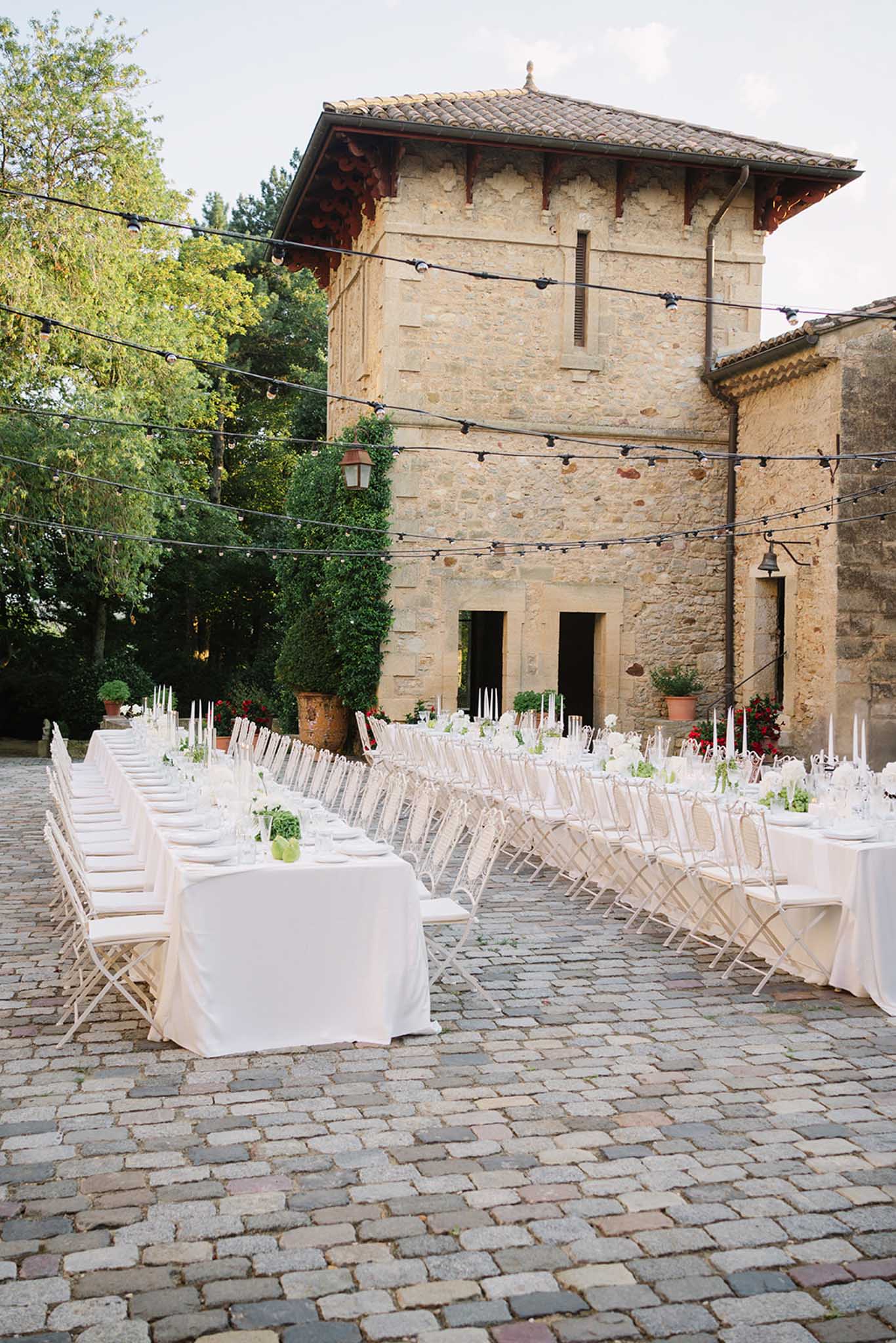 Two long reception tables with white linens and chiavari chairs in stone courtyard, string lights and ivy walls