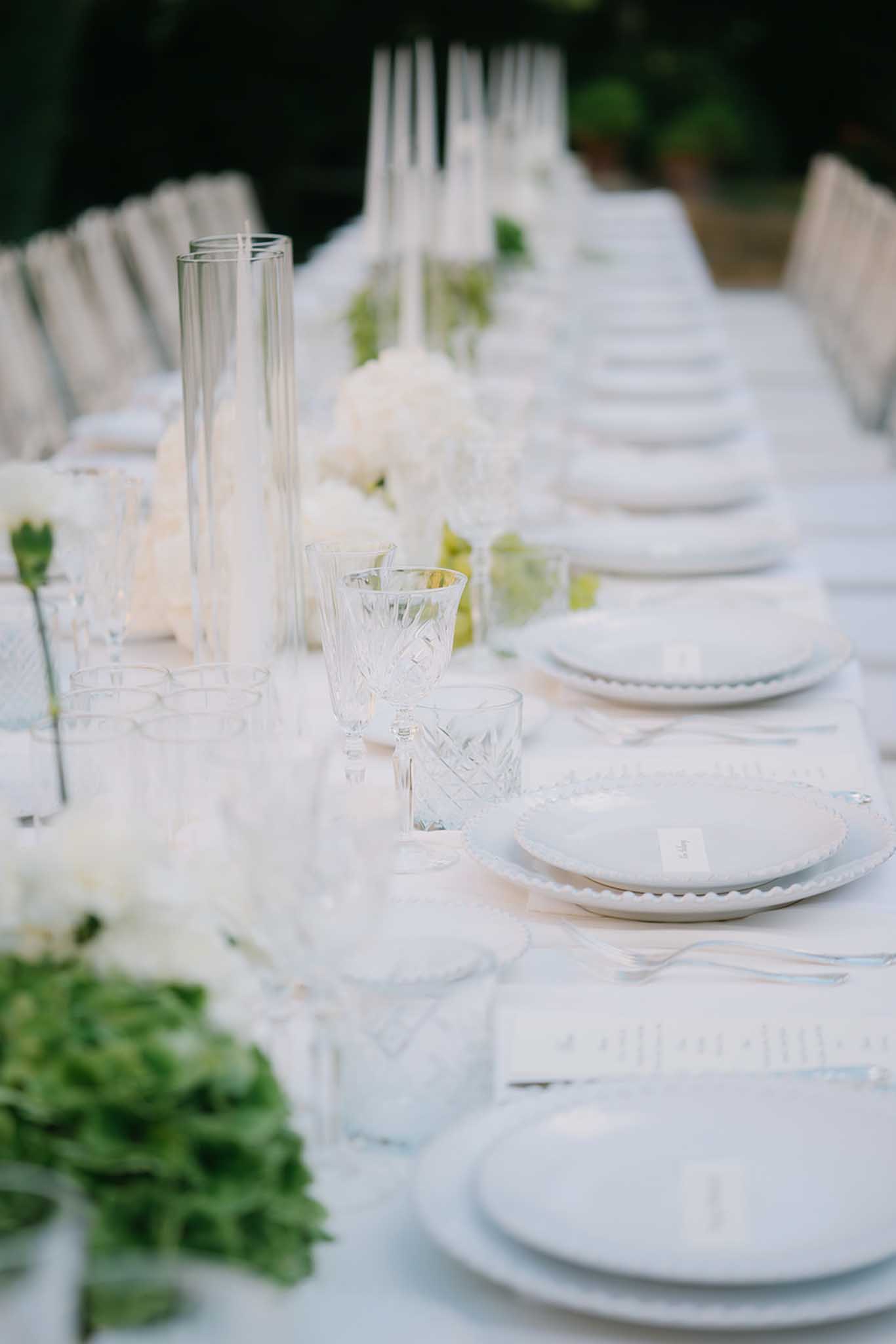 Reception table with white scalloped plates, crystal glassware and white rose hydrangea centrepieces on white linen