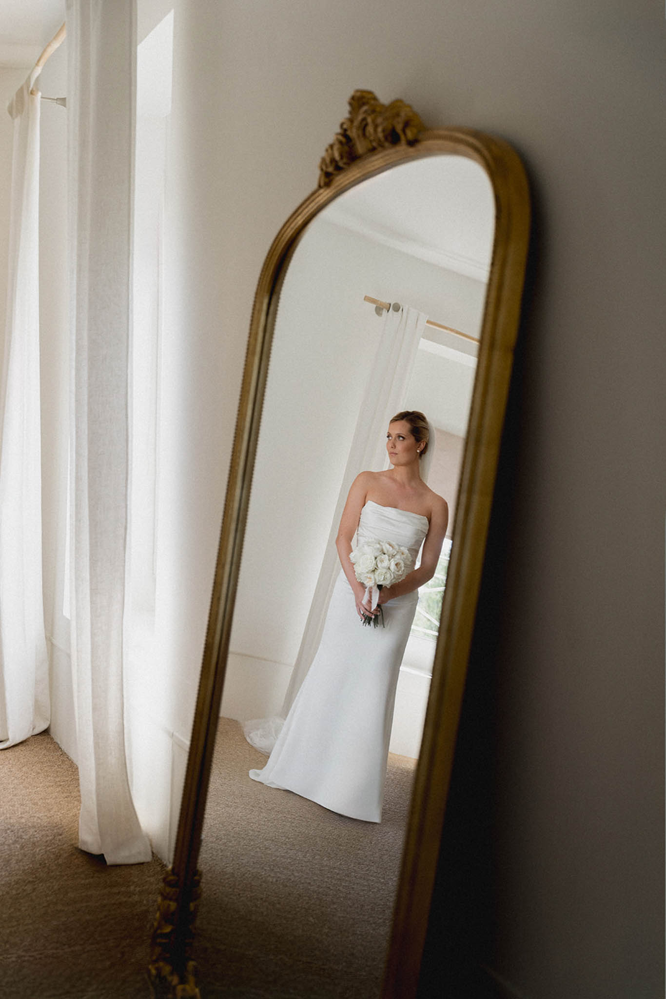 Bride in strapless ivory dress holding white bouquet reflected in ornate gold-framed mirror