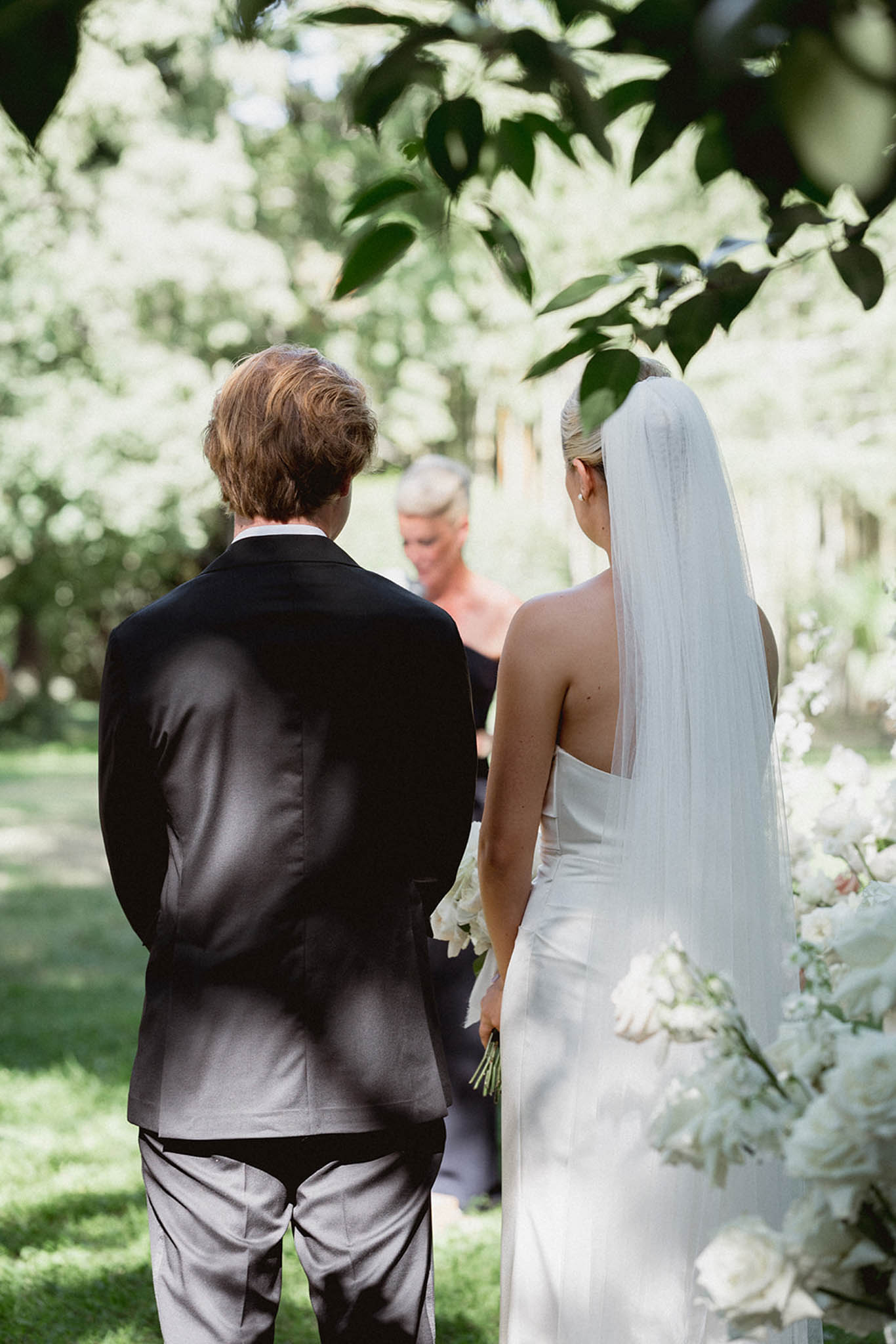 Bride and groom walking down garden aisle from behind, lined with white flowering shrubs and climbing vines