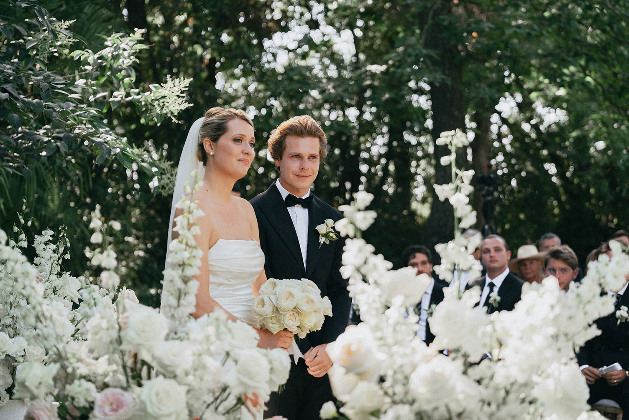 Bride and groom during outdoor ceremony surrounded by white roses, hydrangeas and flowering vines with seated guests behind