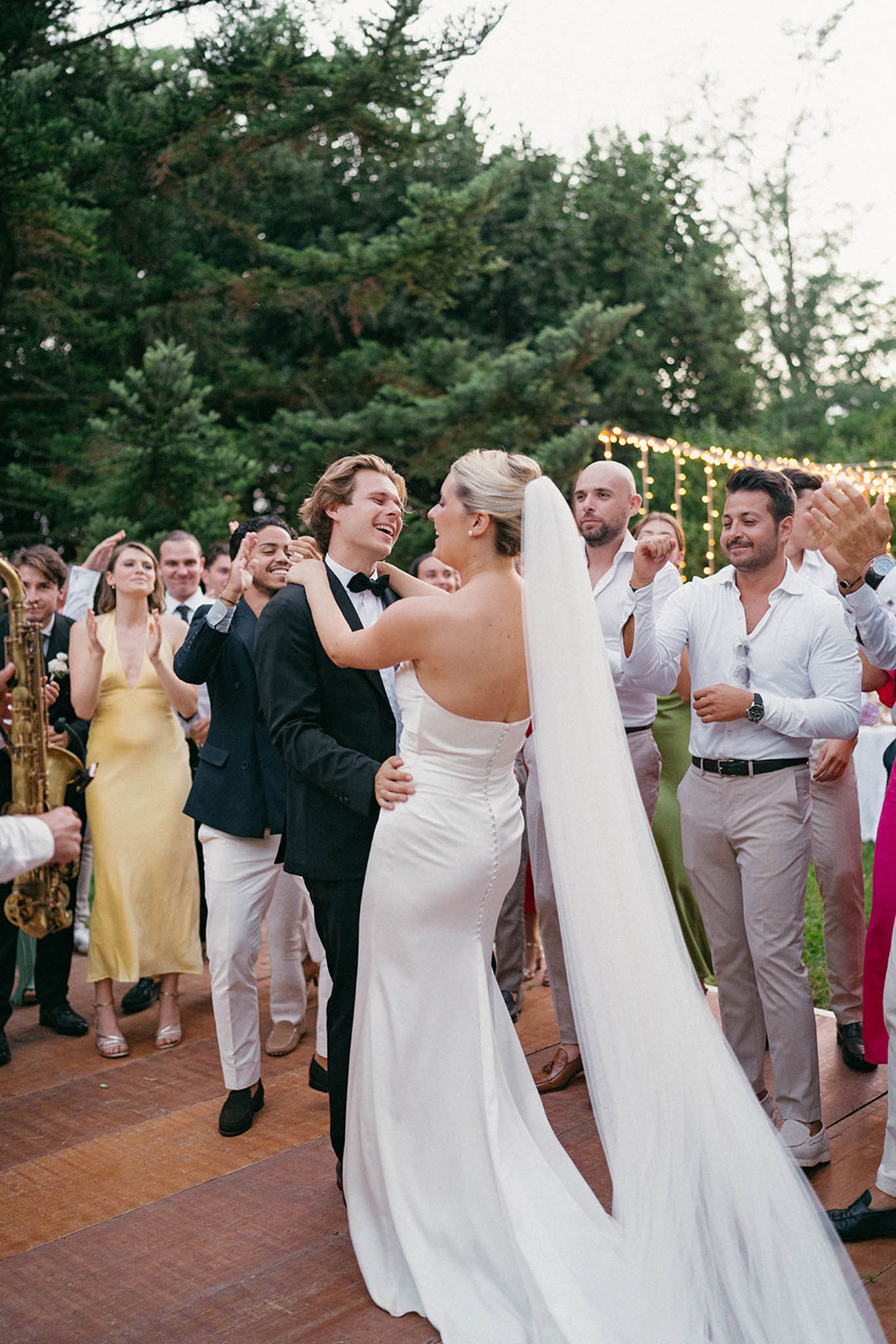 Couple dance outdoors on terrace at night with guests watching, saxophonist performing, and string lights overhead