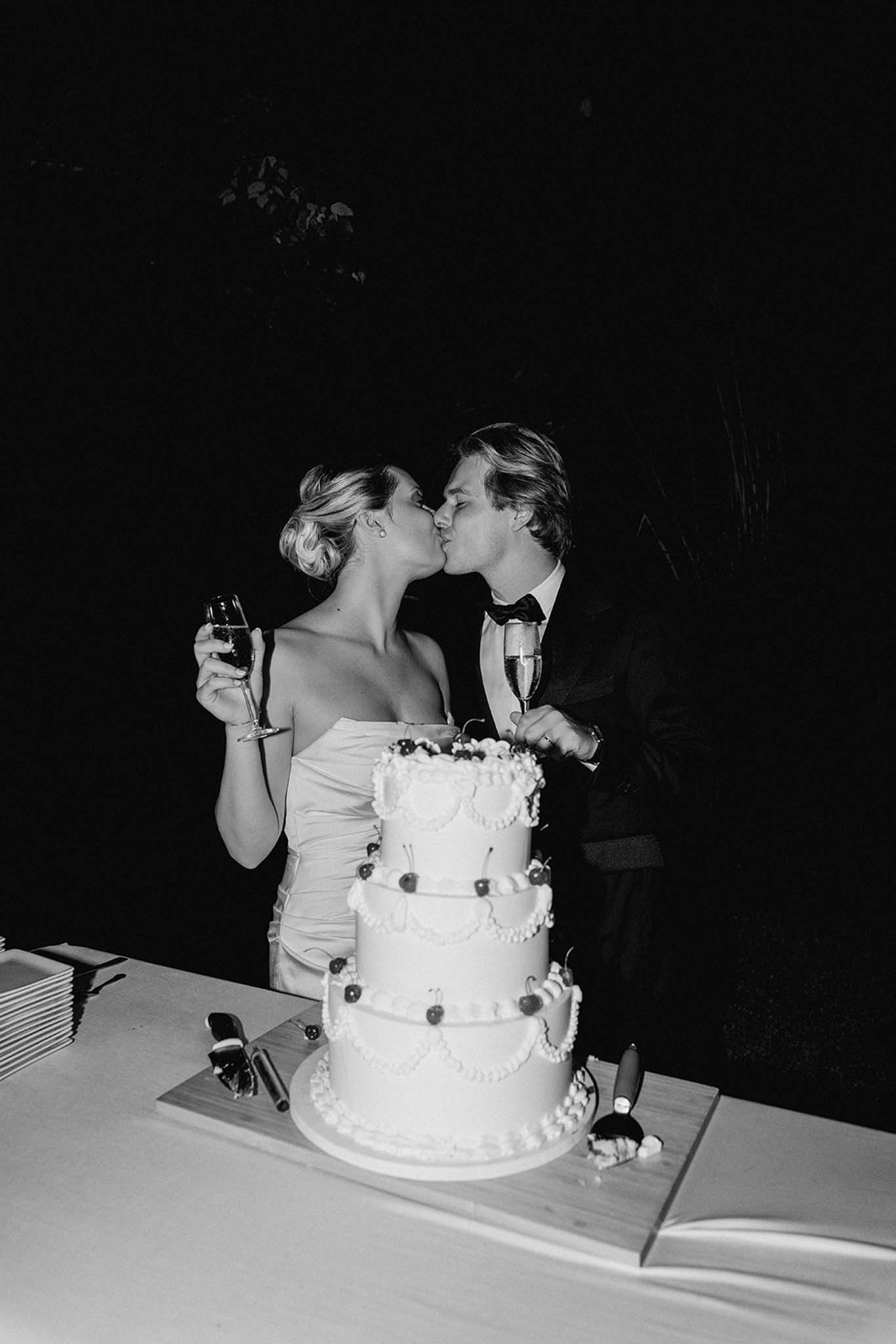 Bride and groom kissing beside three-tiered wedding cake with champagne glasses, black and white photo at indoor reception
