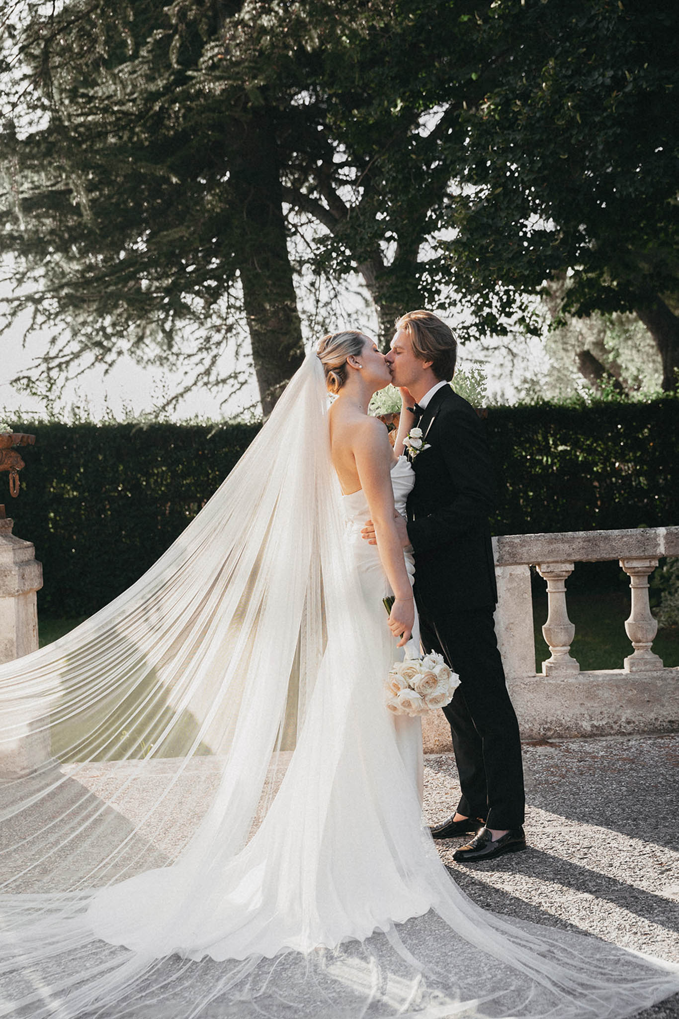 Groom kissing bride's shoulder outdoors in formal garden, bride in strapless gown with long tulle veil trailing