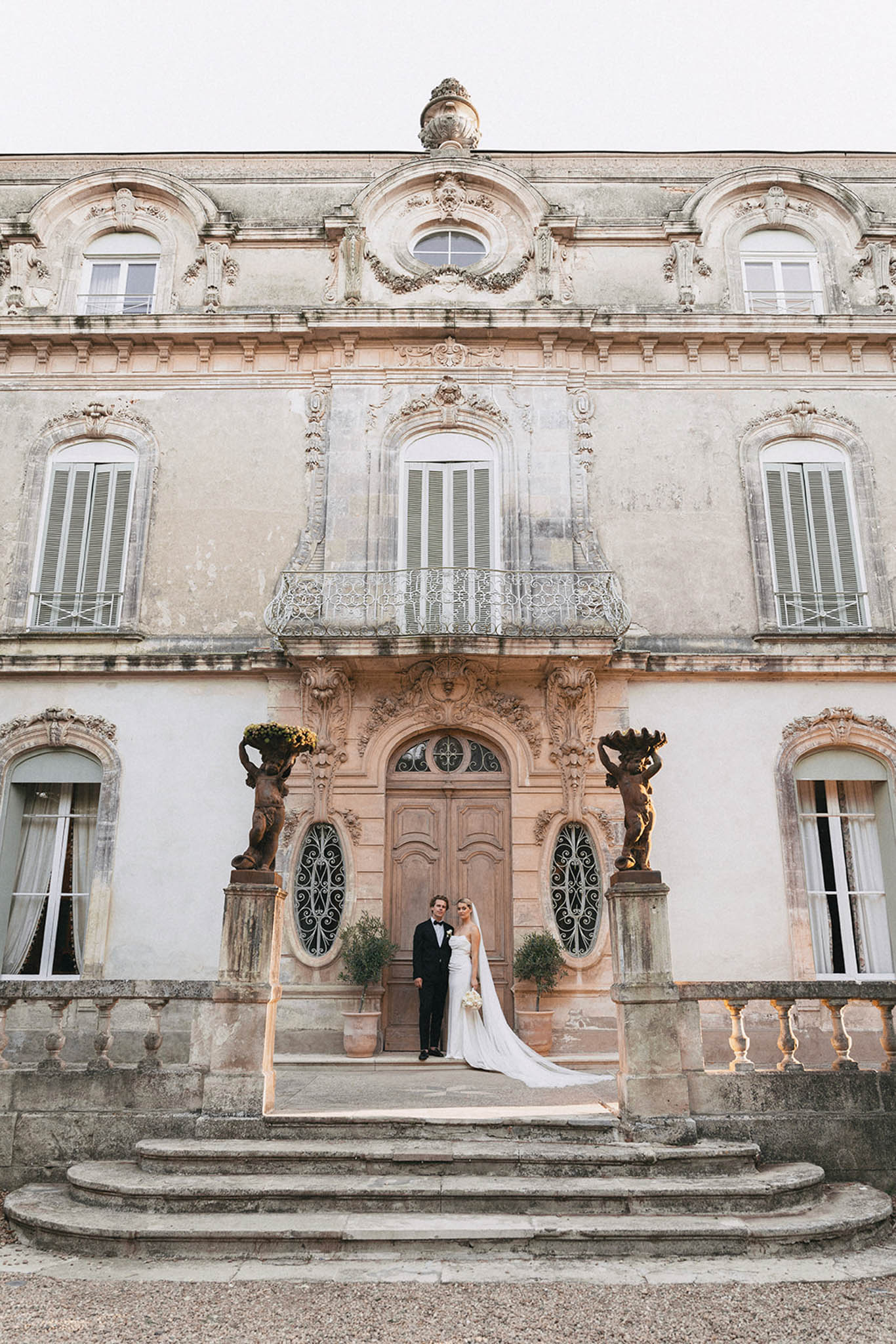Bride and groom on steps of neoclassical stone manor with three stories of arched windows and bronze sculptural urns