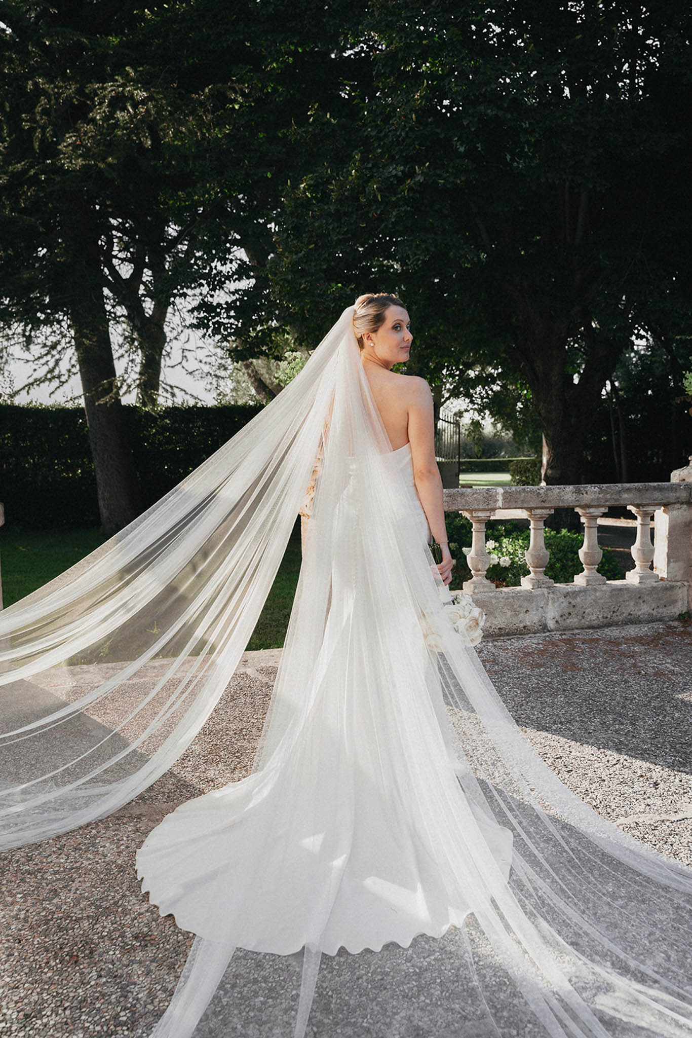 Bride in sleeveless ivory dress with dramatic multi-layered tulle veil sweeping behind her in formal garden