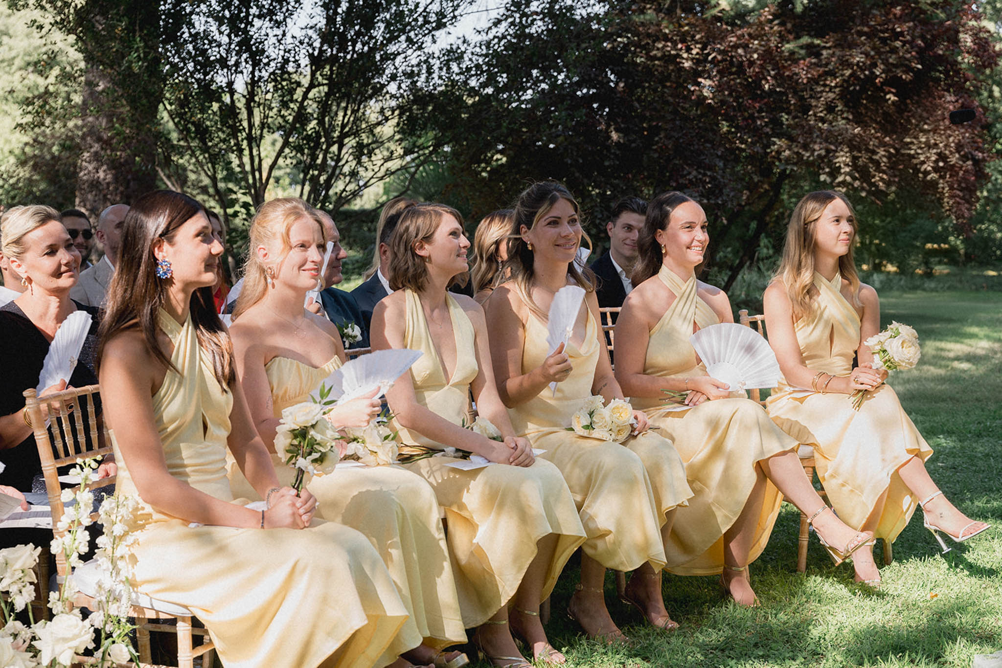 Five bridesmaids in pale yellow dresses holding white bouquets and paper fans seated at garden ceremony