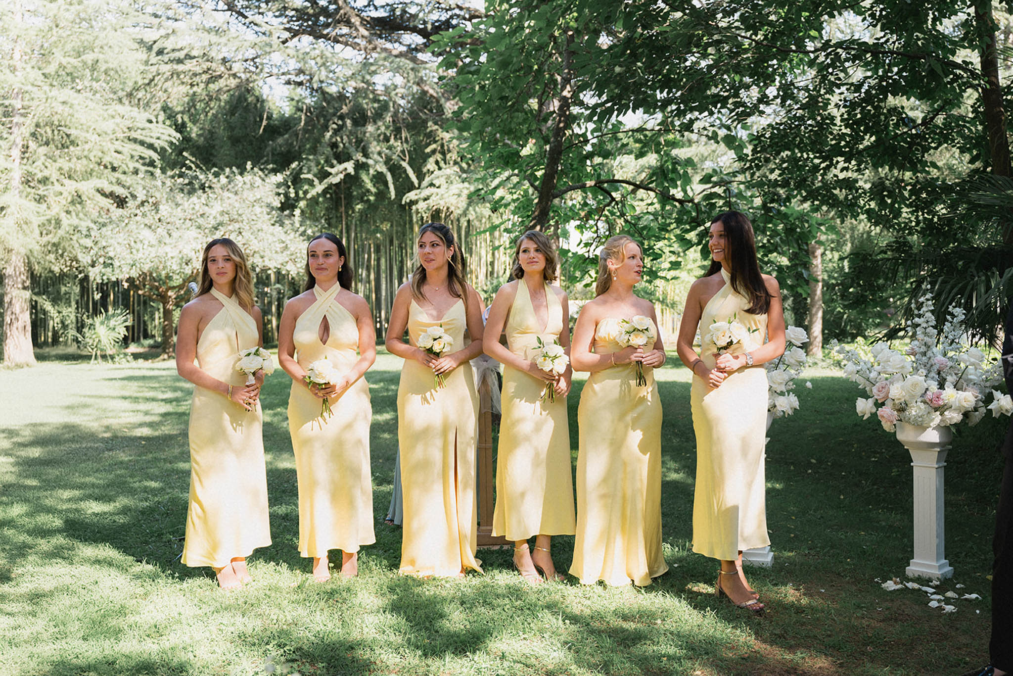 Six bridesmaids in pale yellow dresses holding white bouquets standing on manicured lawn