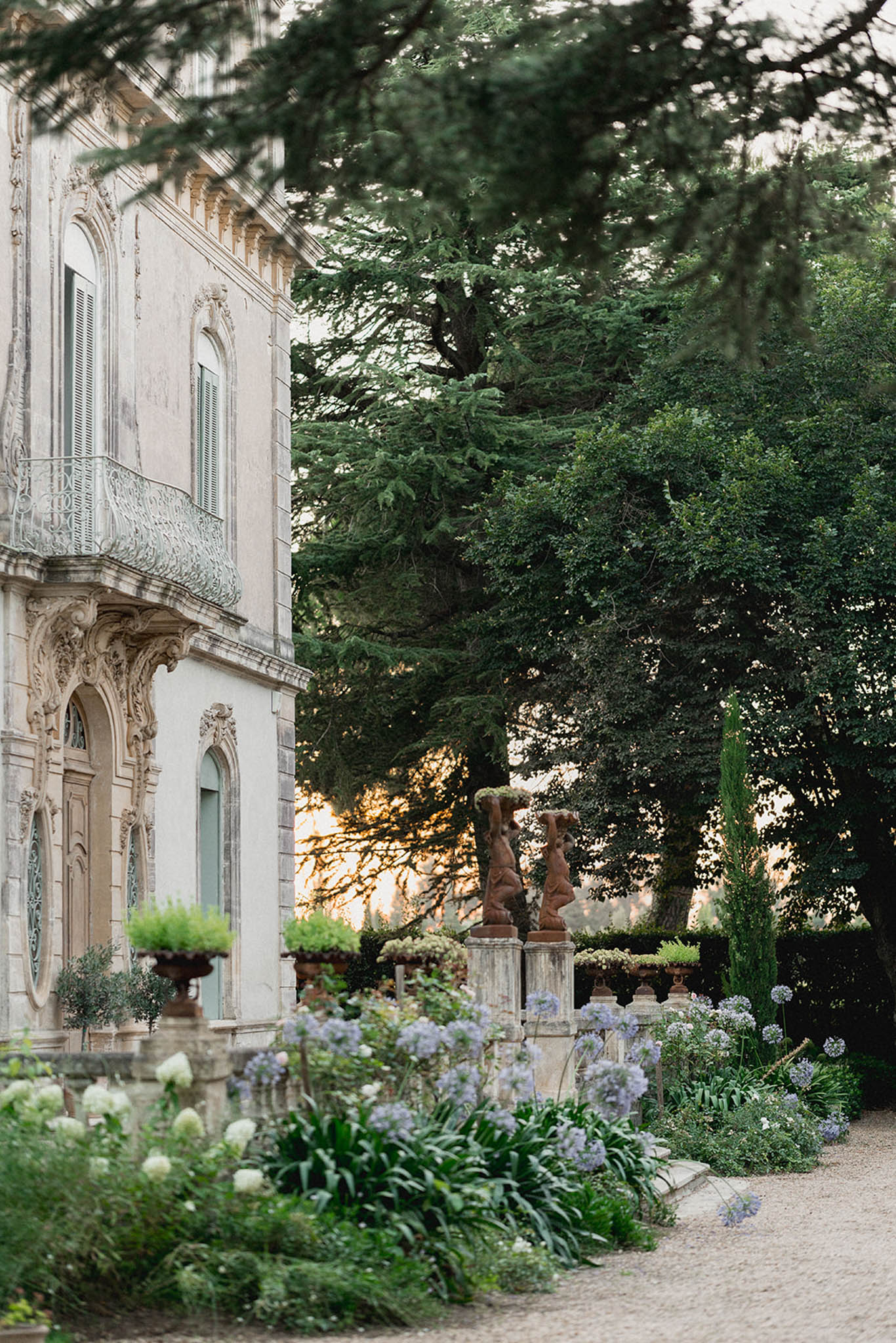 Exterior of a classical French mansion with manicured garden path, blue agapanthus, cypress trees and stone sculptural figures.