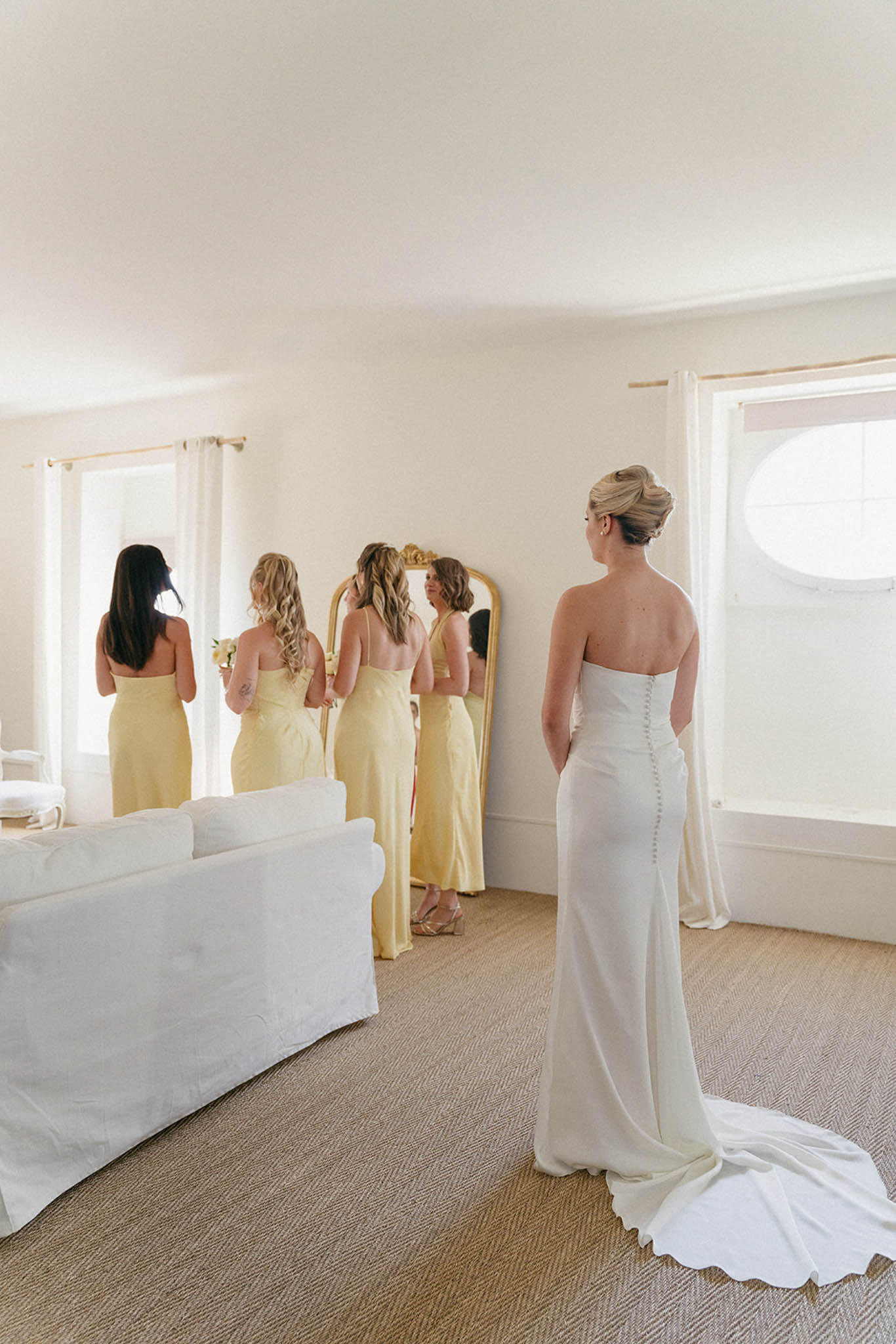 Bride in ivory strapless gown with four bridesmaids in pale yellow dresses during final fitting in bright room