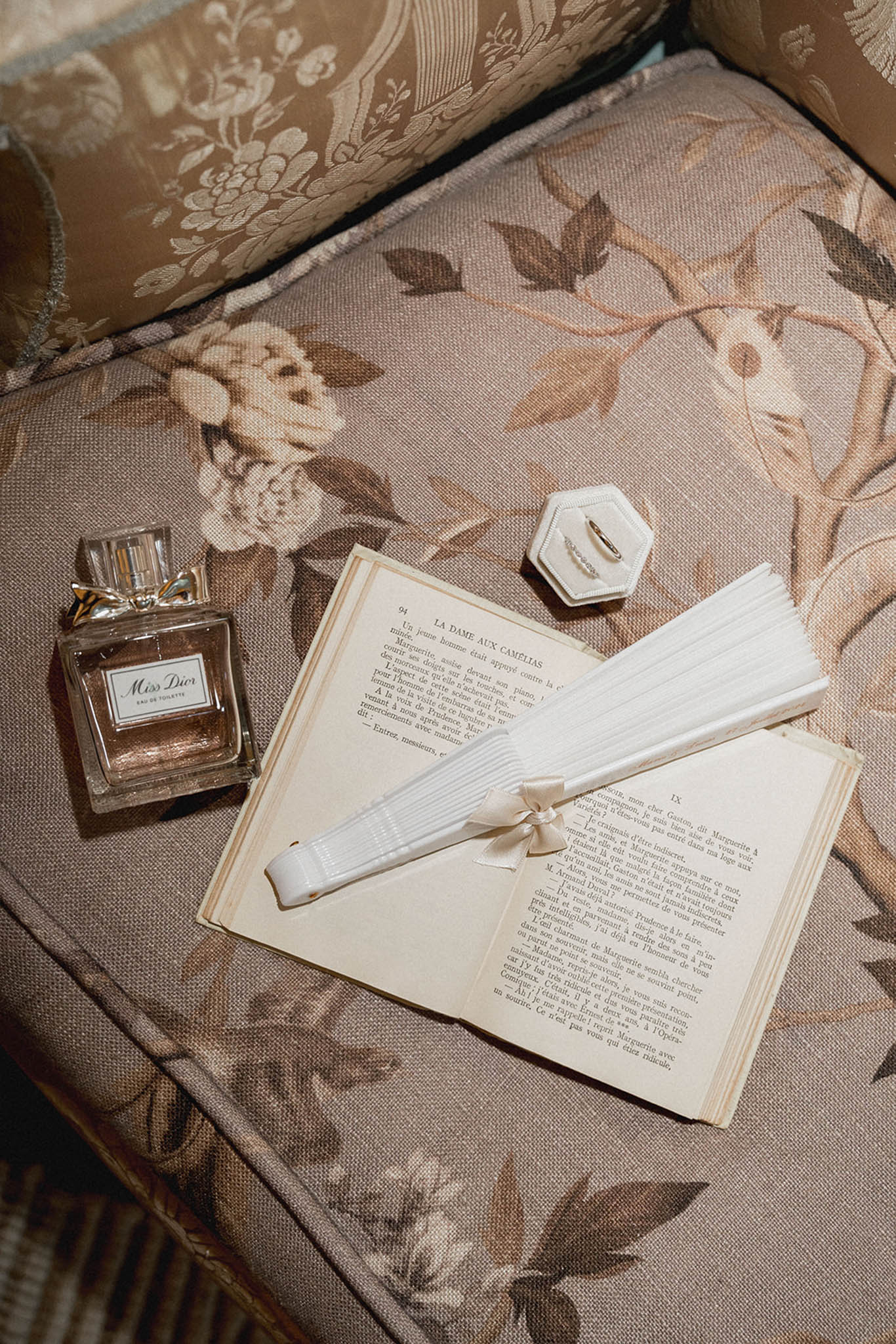 Bridal flat lay with Miss Dior perfume, vintage French novel, silk ribbon, and ring dish on floral fabric