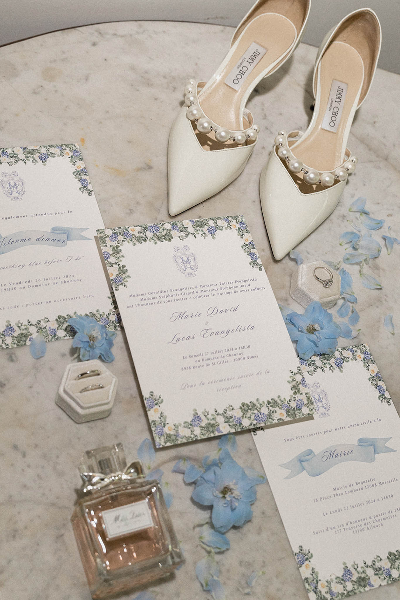 Flat lay of bridal accessories on marble: ivory pearl-embellished heels, periwinkle-bordered wedding invitations, rings and perfume.