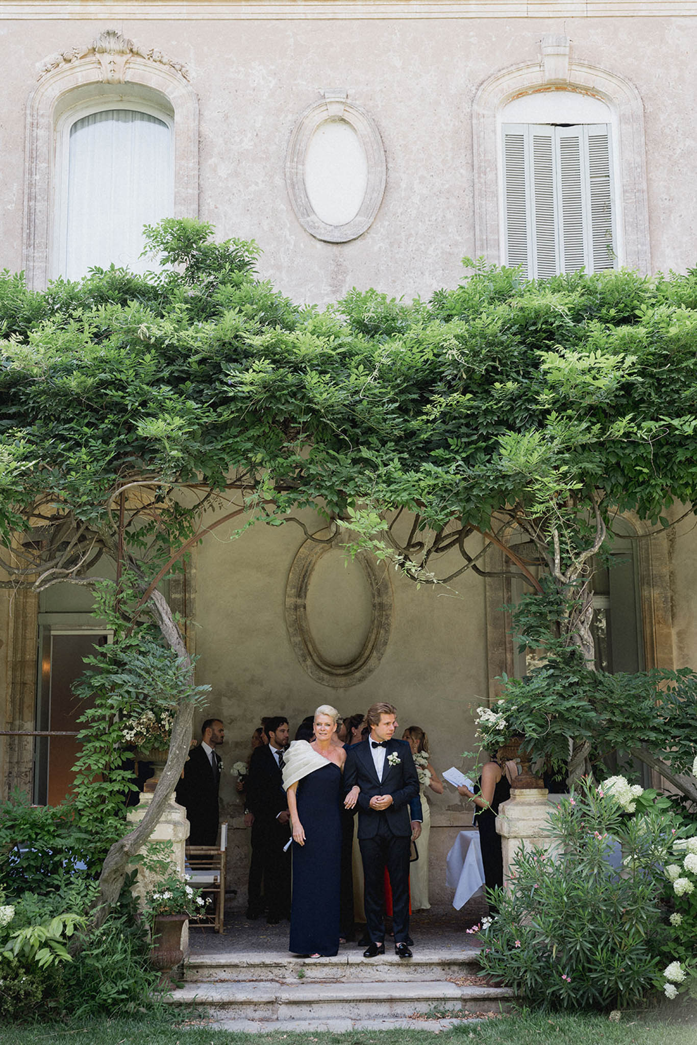 Guests in formal attire mingling near the ivy-covered stone entrance of a historic Italian courtyard during cocktail hour.