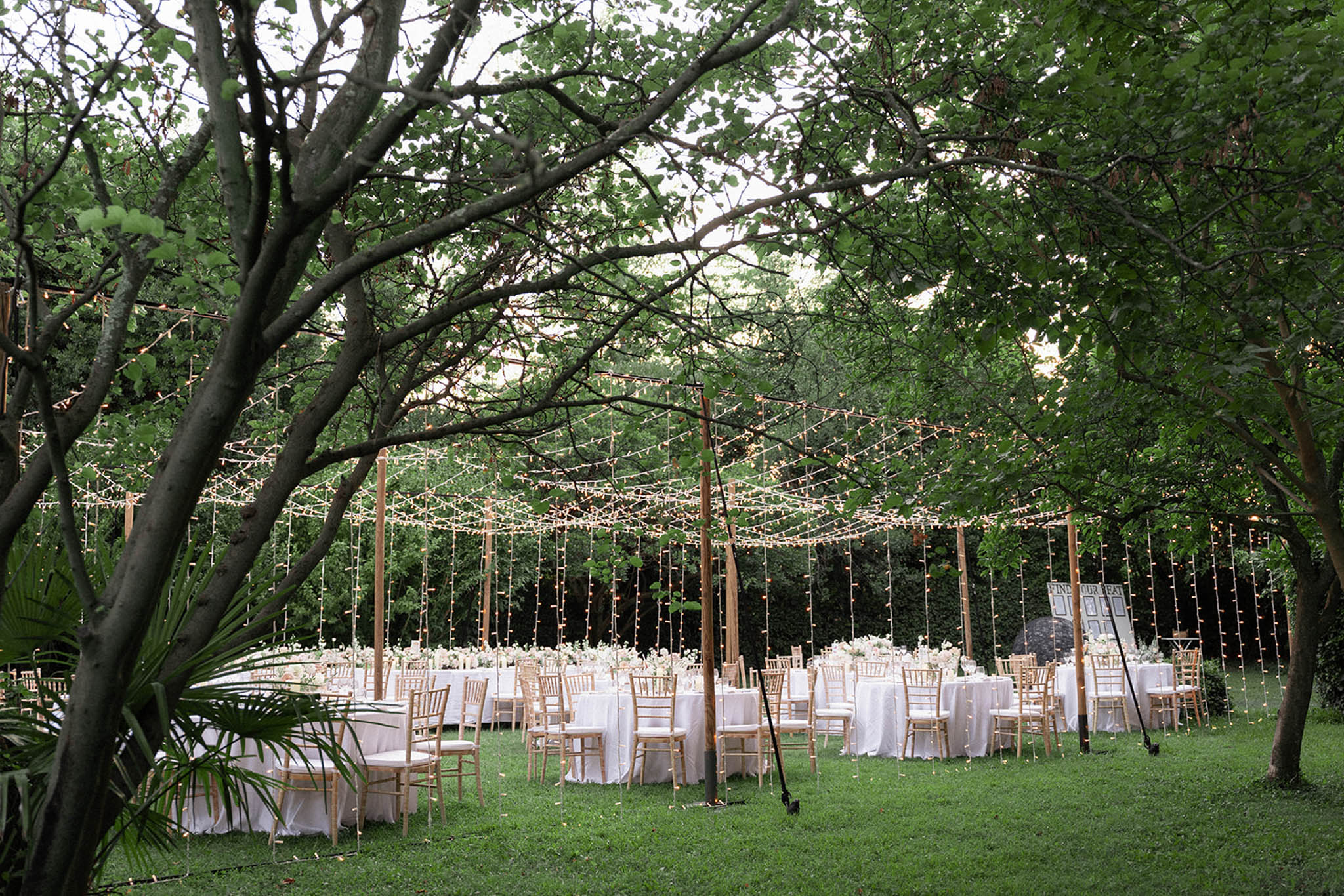 Outdoor reception with round tables and string lights beneath spreading trees in garden