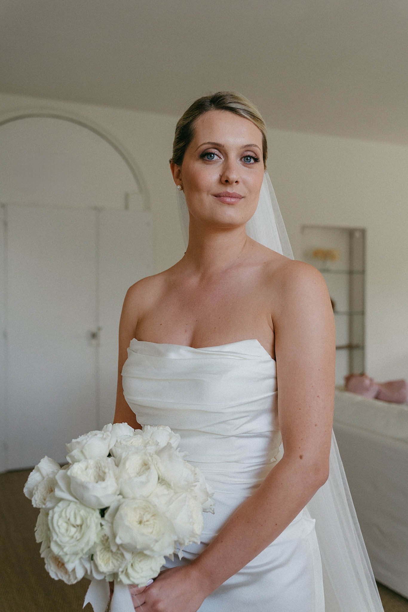 Bride in strapless ruched gown with simple veil posing indoors against neutral walls