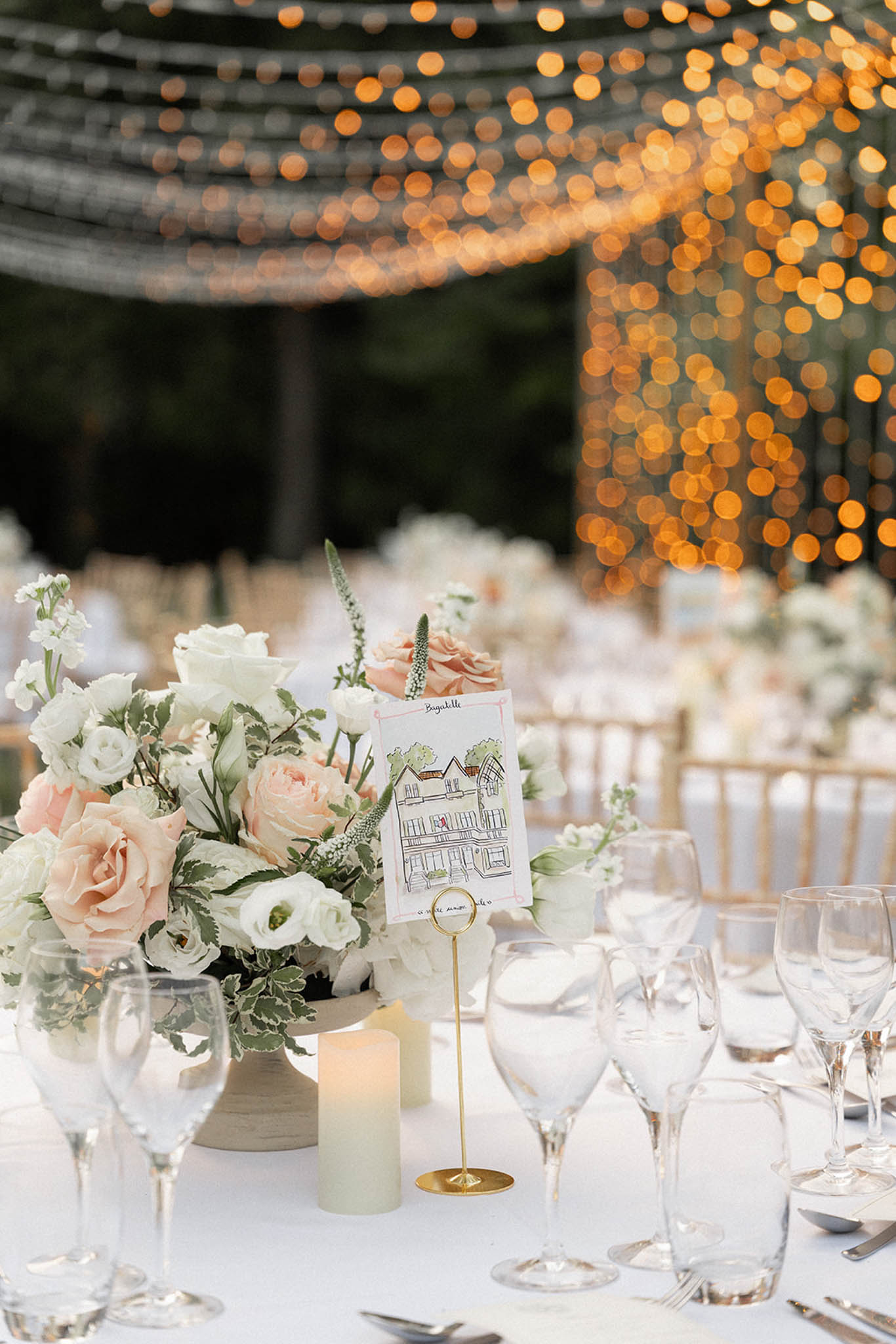 Reception table at twilight with ivory and blush floral centerpiece in grey vase, brass place card holder, and golden string lights overhead
