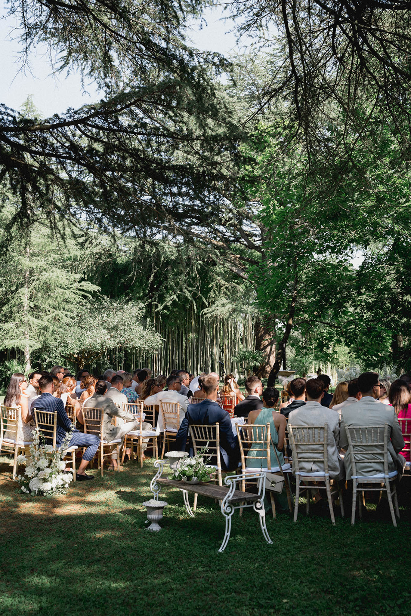 Outdoor ceremony under tree canopy with macramé backdrop, 60-80 guests on Chiavari chairs and white florals