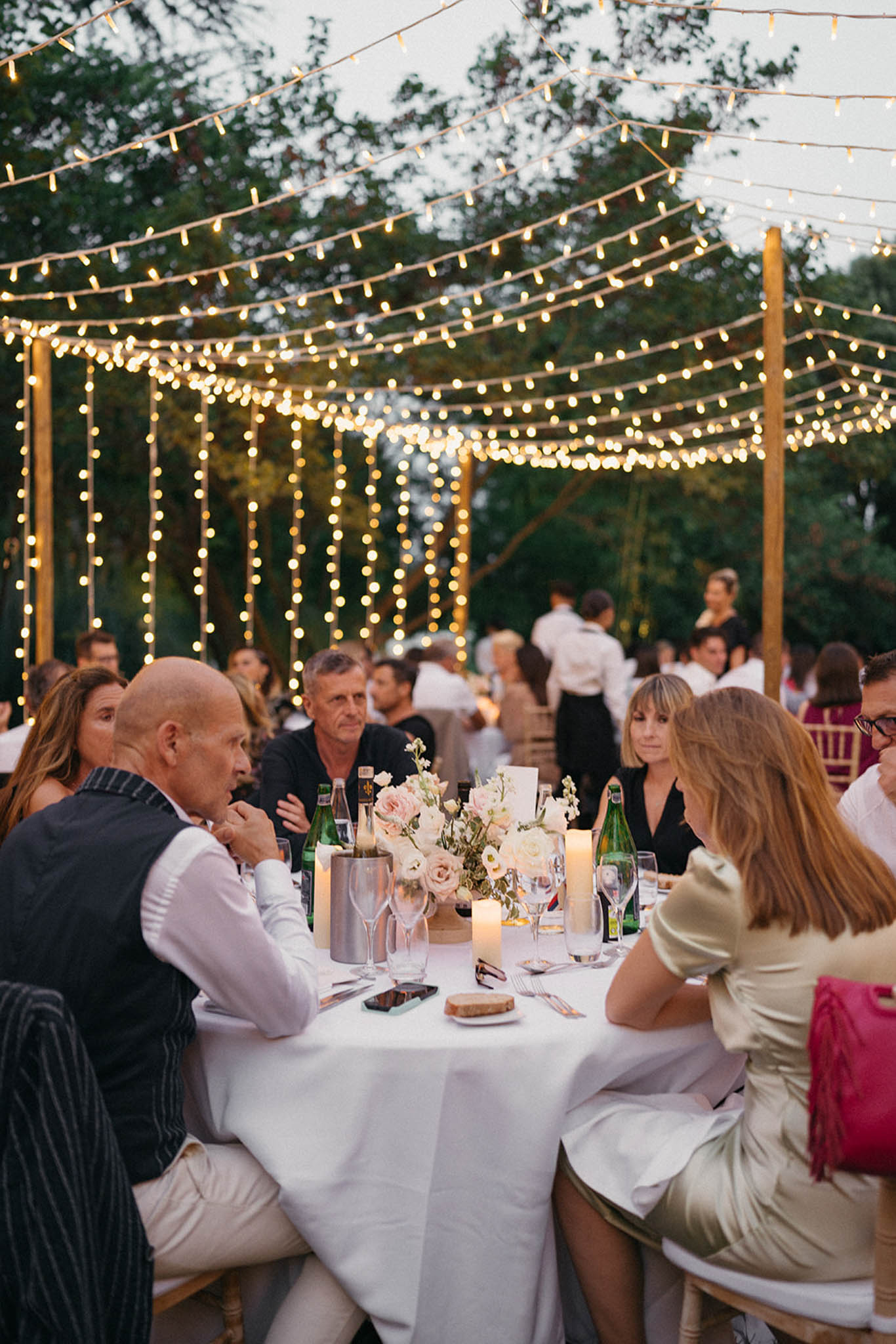 Guests seated at round tables under warm string light canopy in outdoor garden reception