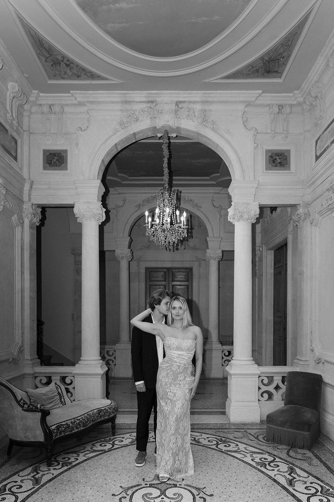 Black and white portrait of bride and groom in ornate neoclassical hall with columns and crystal chandelier