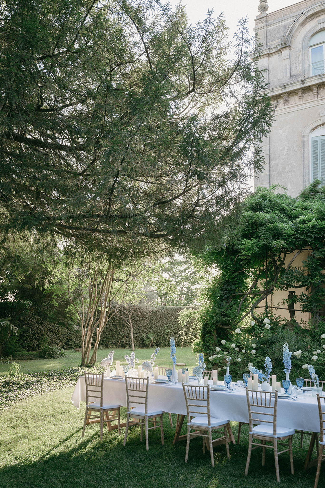 Garden reception with round tables, white linens, Chiavari chairs, blue delphiniums and white hydrangeas, stone building