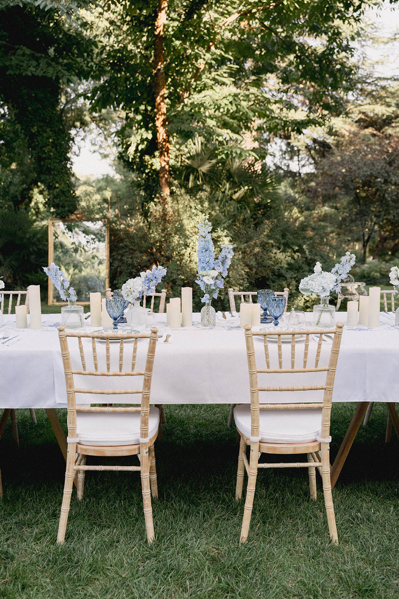 Long outdoor reception tables with white lilac arrangements in blue glass vases, ivory pillar candles and wooden chiavari chairs.