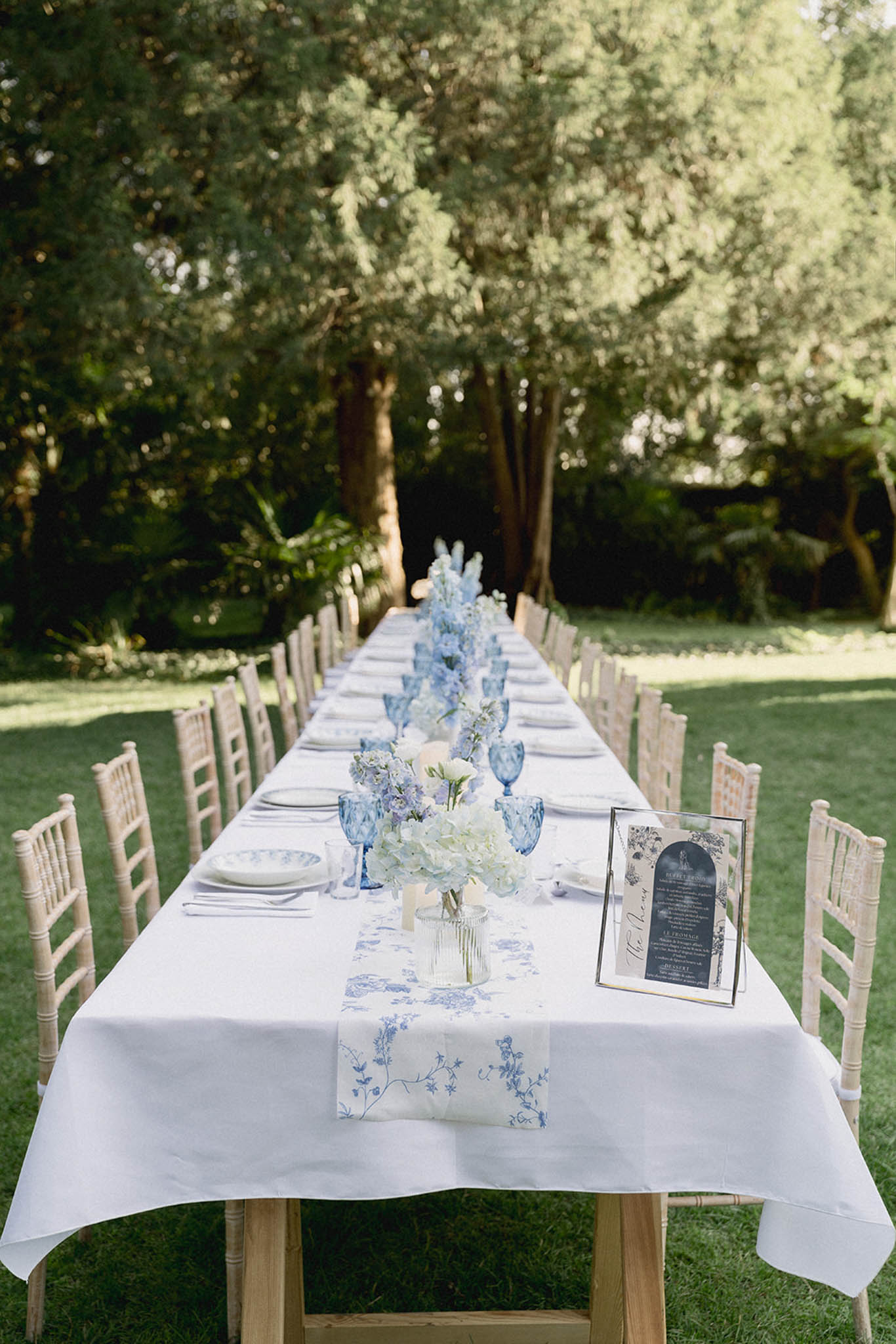 Long outdoor reception table with blue and white patterned runner, white hydrangea centerpieces, and blue goblets