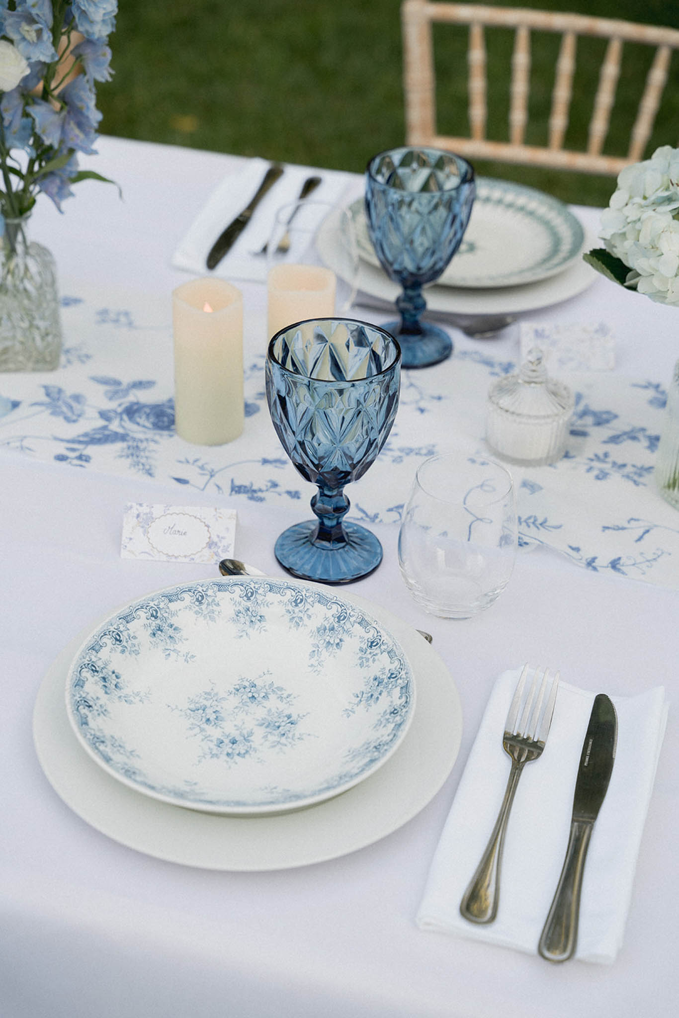 Close-up of outdoor reception place setting with blue botanical-print plates, textured blue glass goblets and white hydrangea centerpiece.