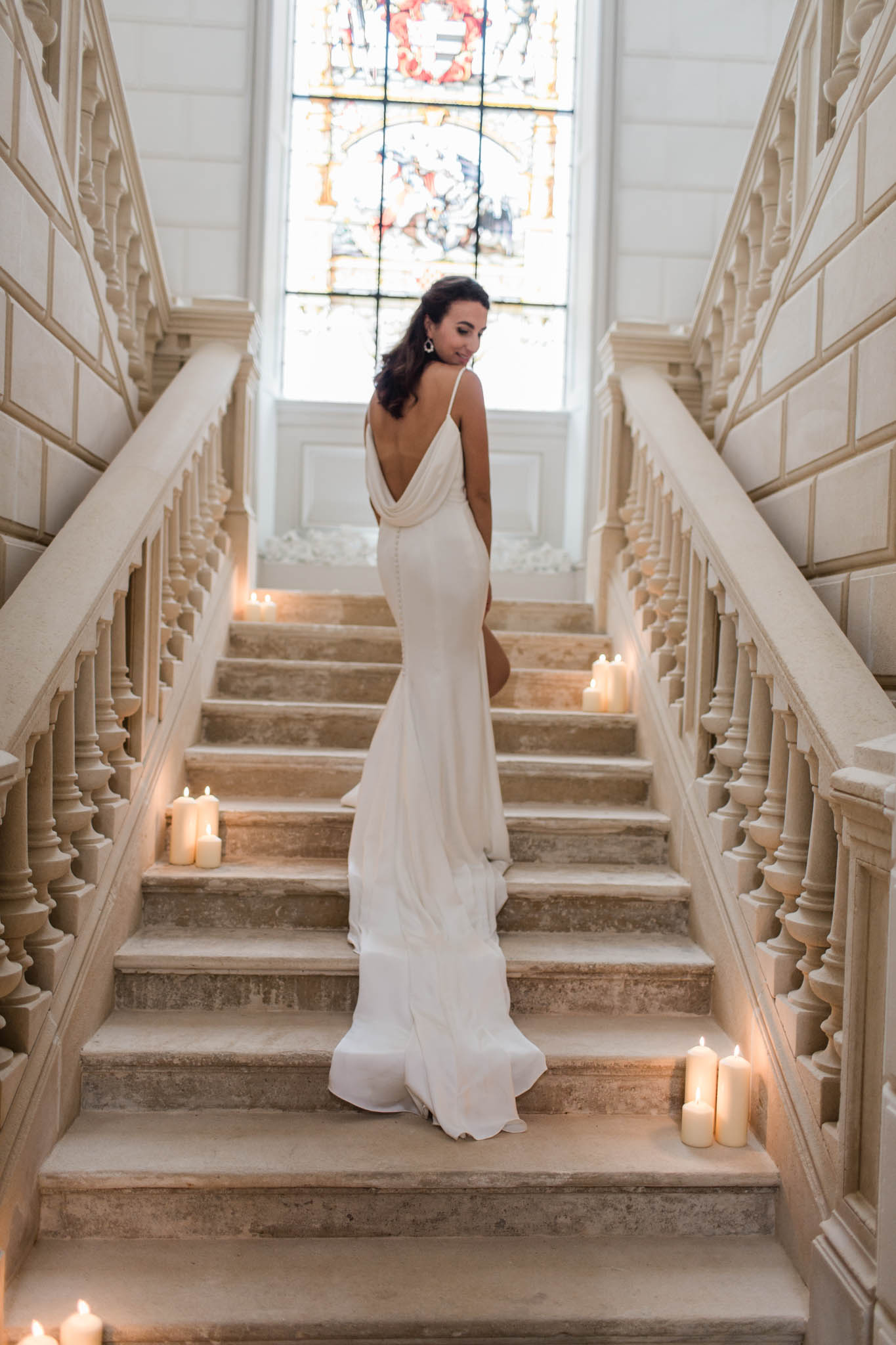 Bride in fitted ivory crepe gown with deep V-back descending grand staircase with candles, stained-glass window above