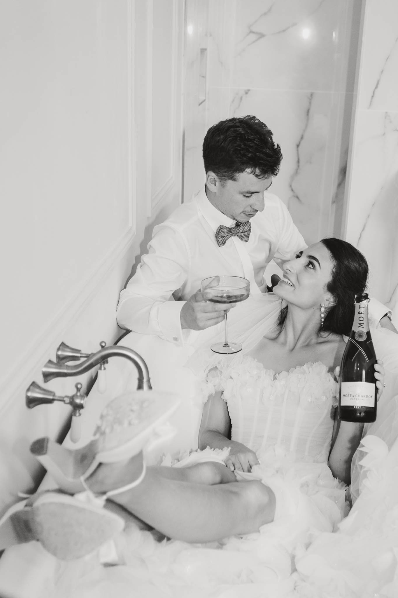 Bride and groom sitting together in bathtub holding wine glasses, black and white photo at Château de Londigny