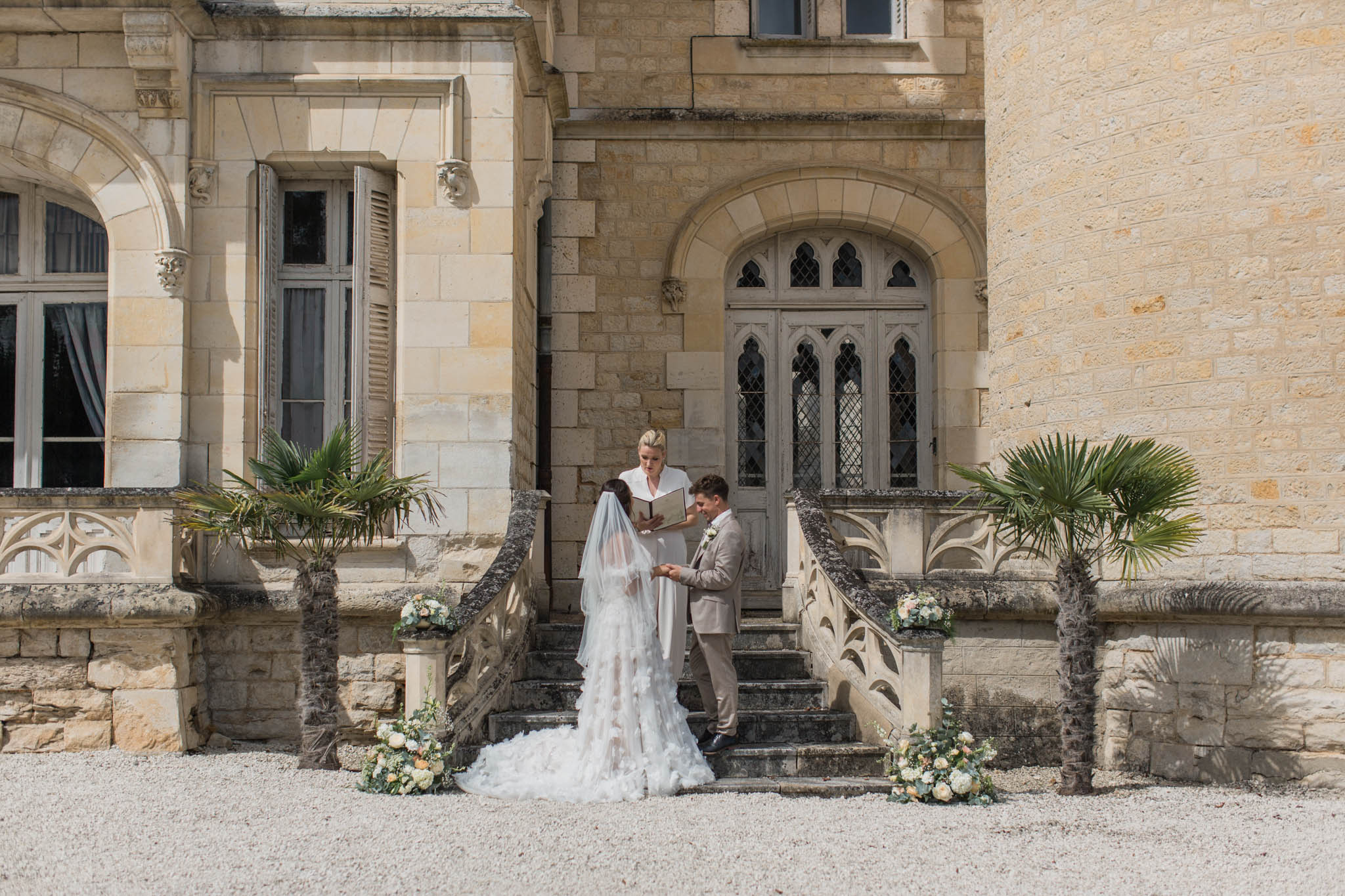 Wedding ceremony on stone manor steps, bride in ivory lace gown, groom in grey suit, officiant in white, potted palms