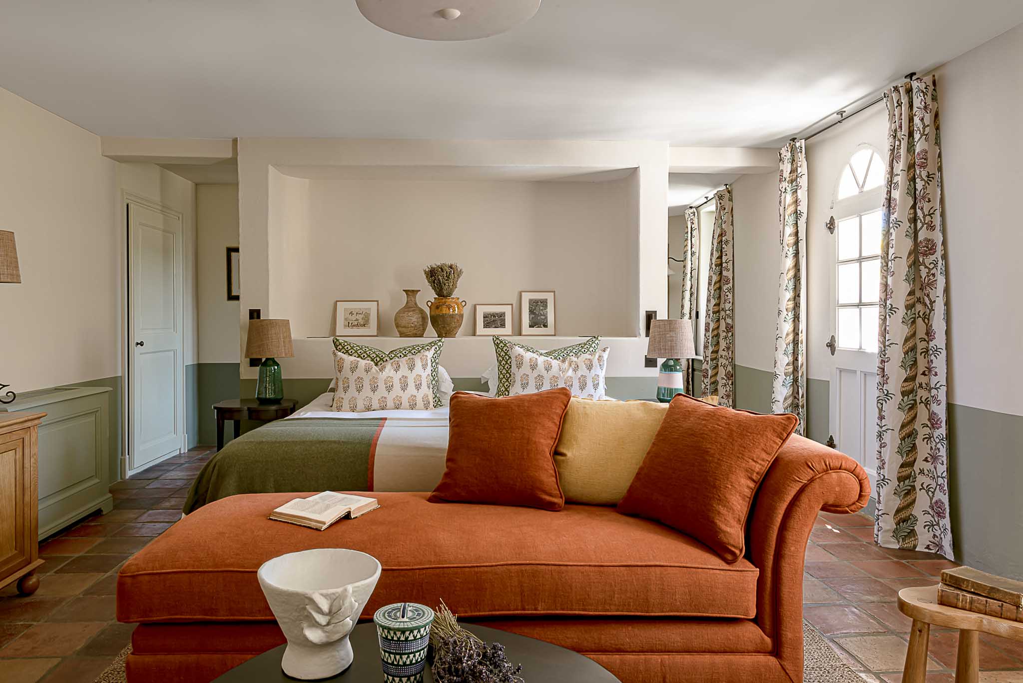 Guest bedroom with white linens, terracotta chaise longue, sage wainscoting, and ceramic decor in French country property