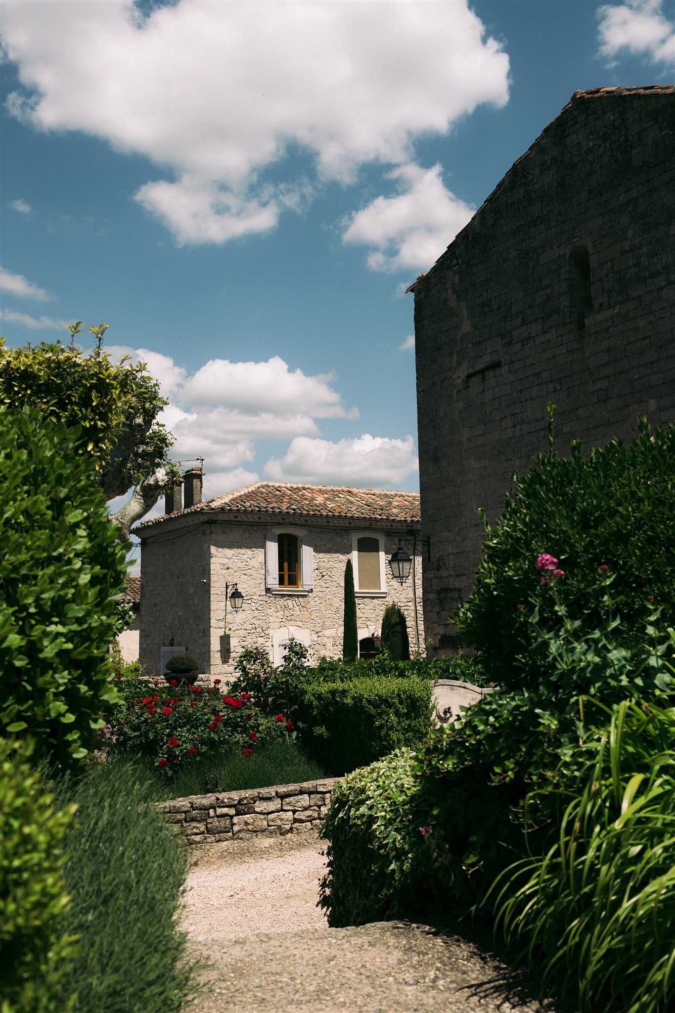 Historic stone venue with clay tile roof and tower, garden pathway with red roses, cypress trees, blue sky above