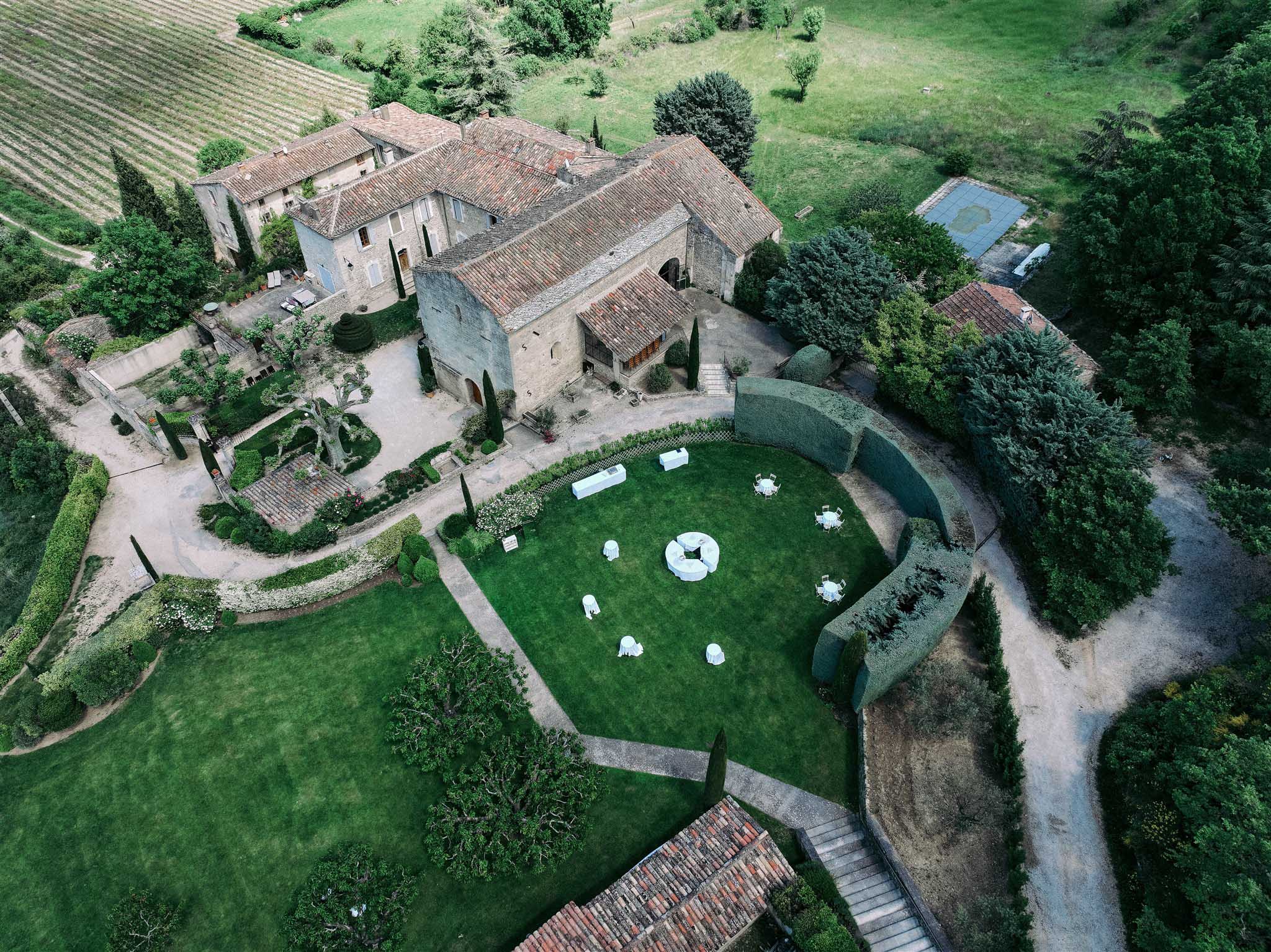 Aerial view of stone villa wedding venue with cocktail tables on lawn, surrounded by cypress trees and vineyard hills