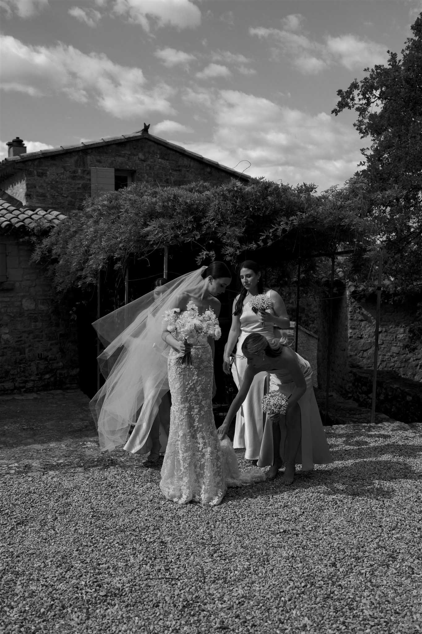 Black and white portrait of bride in lace gown with two bridesmaids in a stone courtyard with olive trees